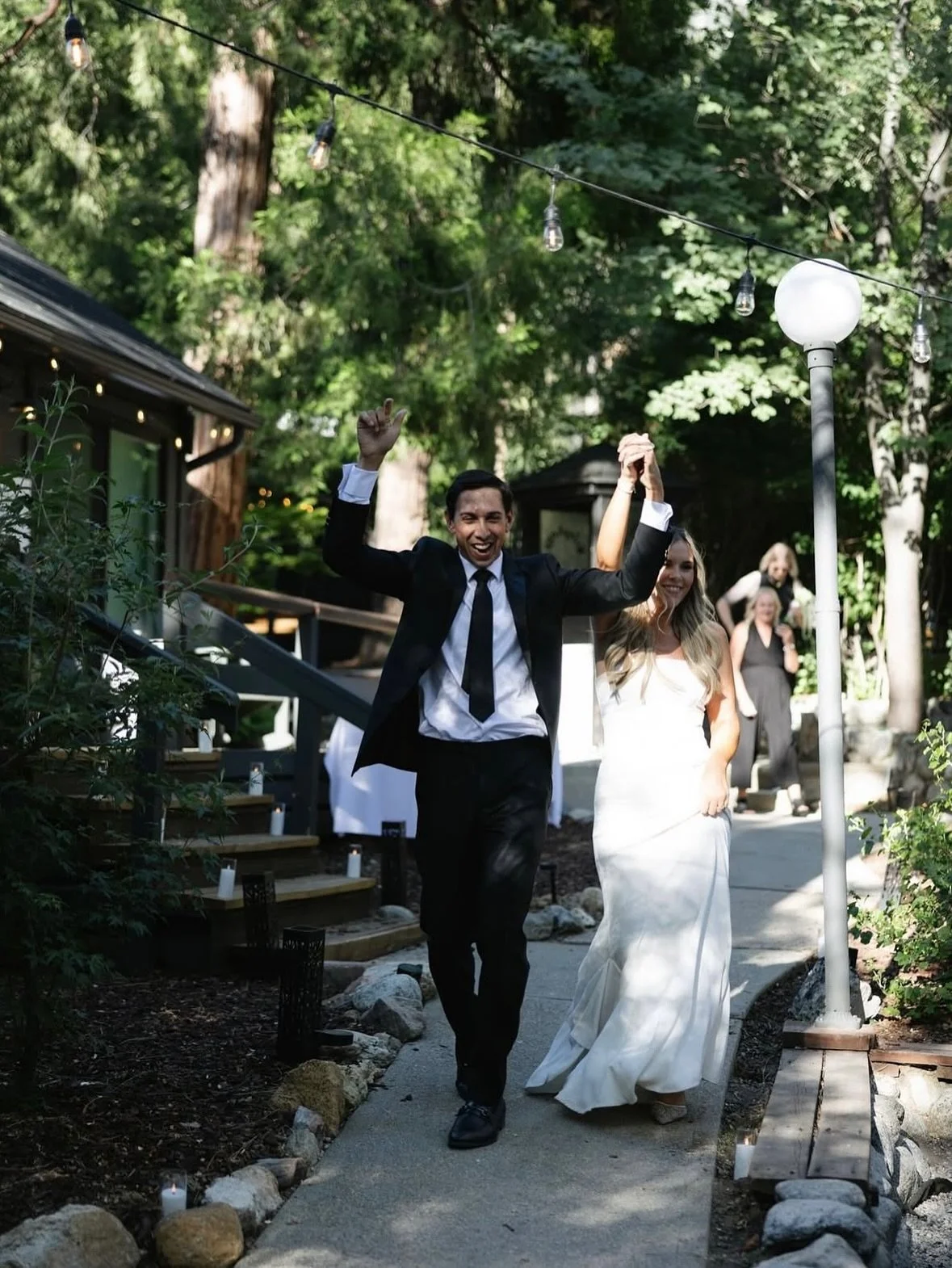 weddings at The Pinetree Hotel . A newlywed couple walking down a garden pathway, celebrating, with a man in a black tuxedo and woman in a white wedding dress, surrounded by greenery and outdoor string lights.