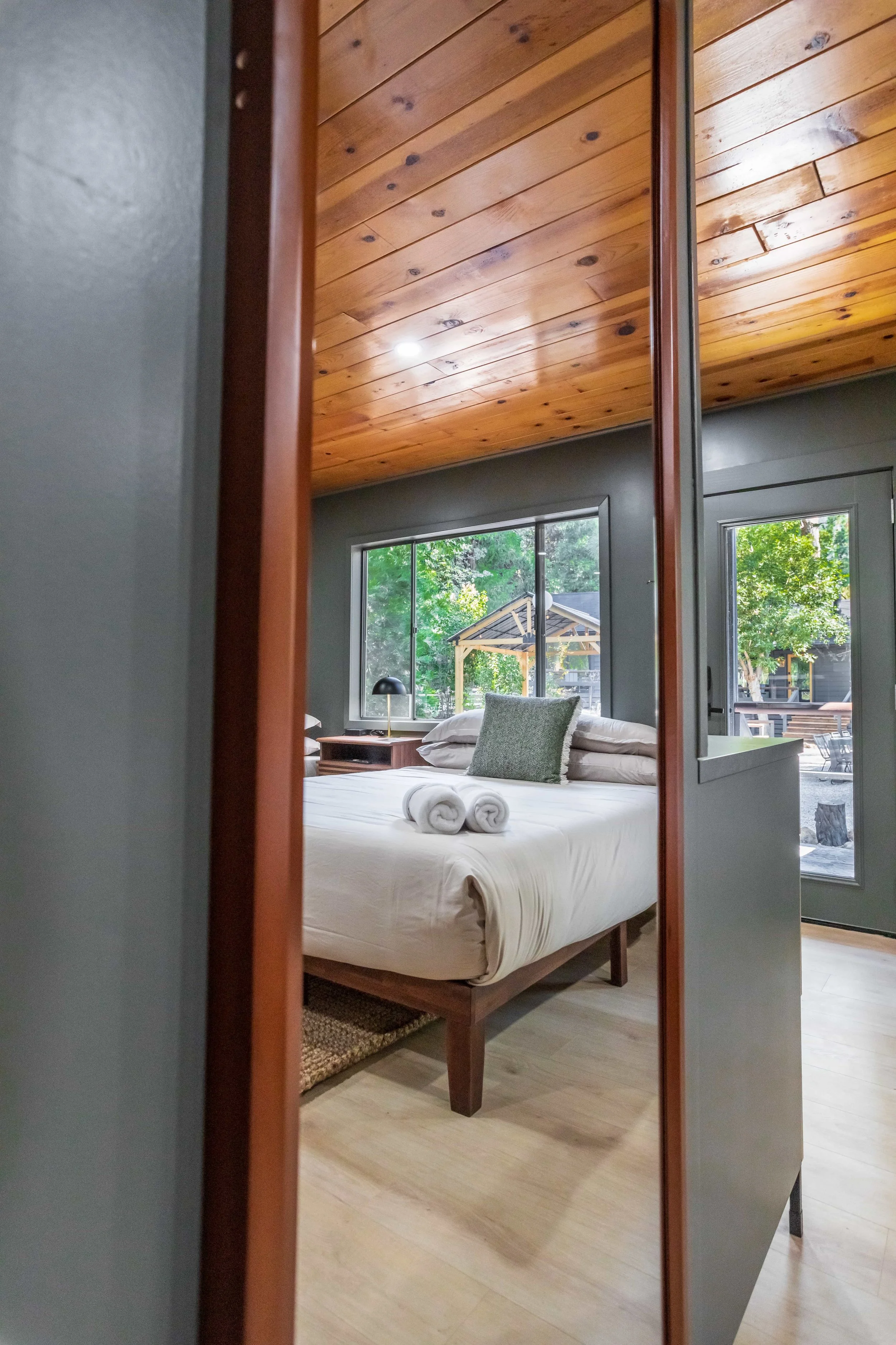 A cozy bedroom with wooden ceiling, large windows, a bed with rolled towels, pillow, and a view of trees outside.