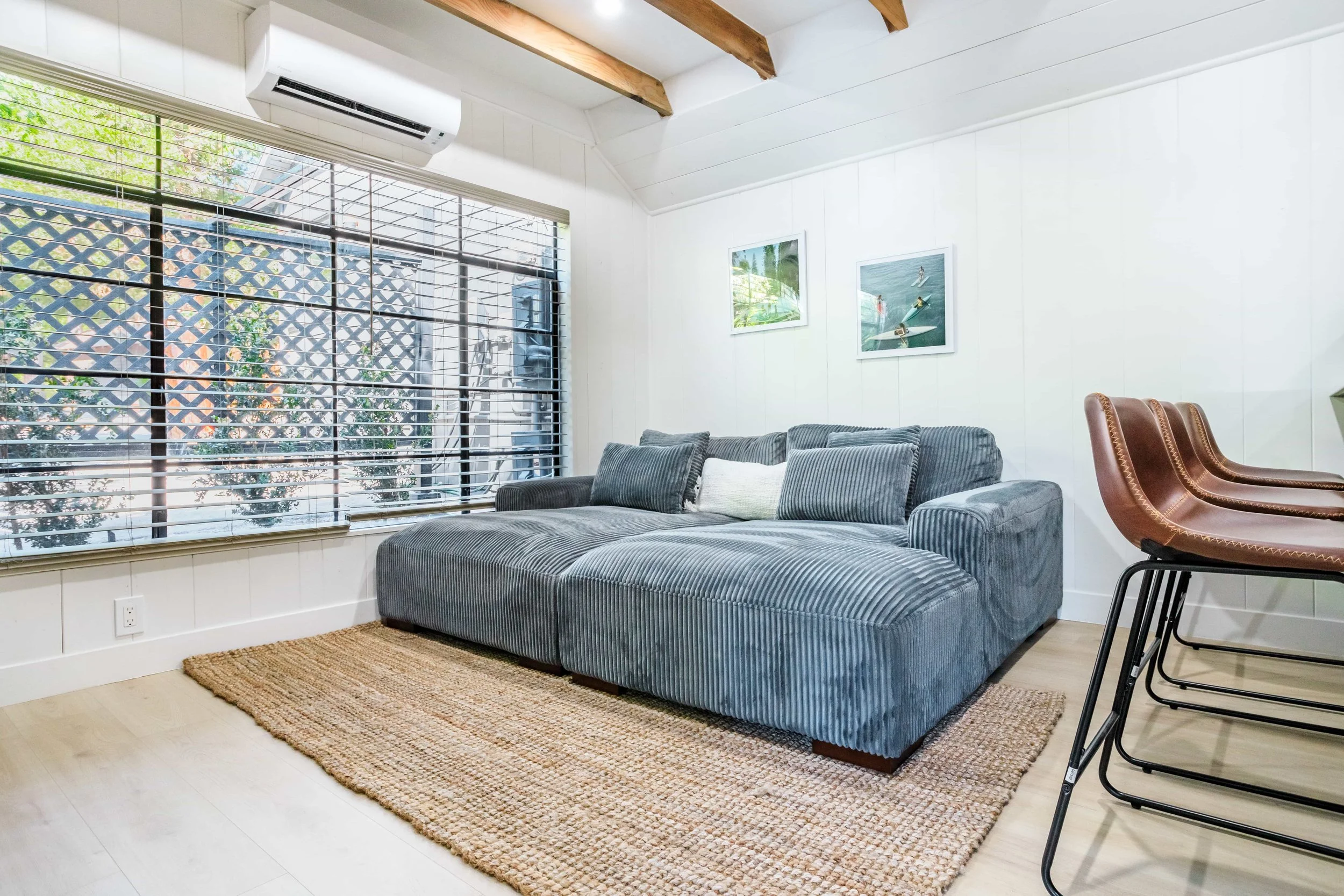 Living room with large window, grey striped sectional sofa on a woven area rug, and three brown leather chairs stacked at the side. The walls are white with two framed ocean photos, and air conditioning unit mounted above the window.