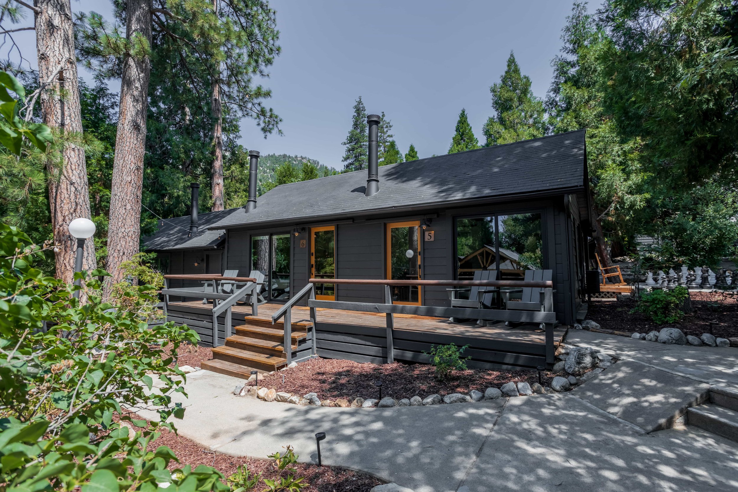 A black wooden cabin with a small front porch, surrounded by tall trees and a landscaped yard, with a winding concrete pathway leading to the porch stairs.