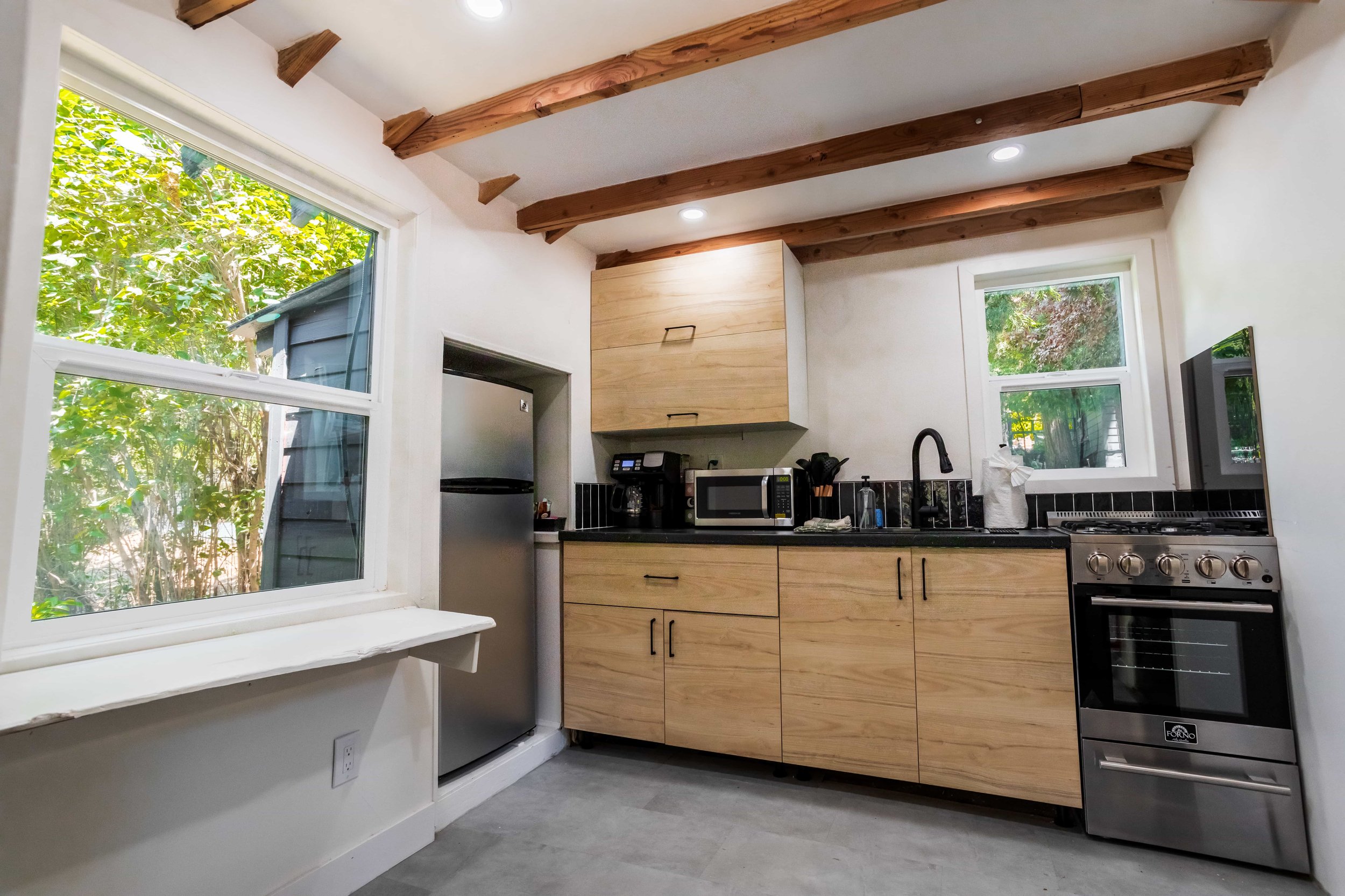 Modern kitchen with wooden cabinets, black countertops, stainless steel refrigerator and stove, black sink and fixtures, microwave, coffee maker, and large windows showing greenery outside.