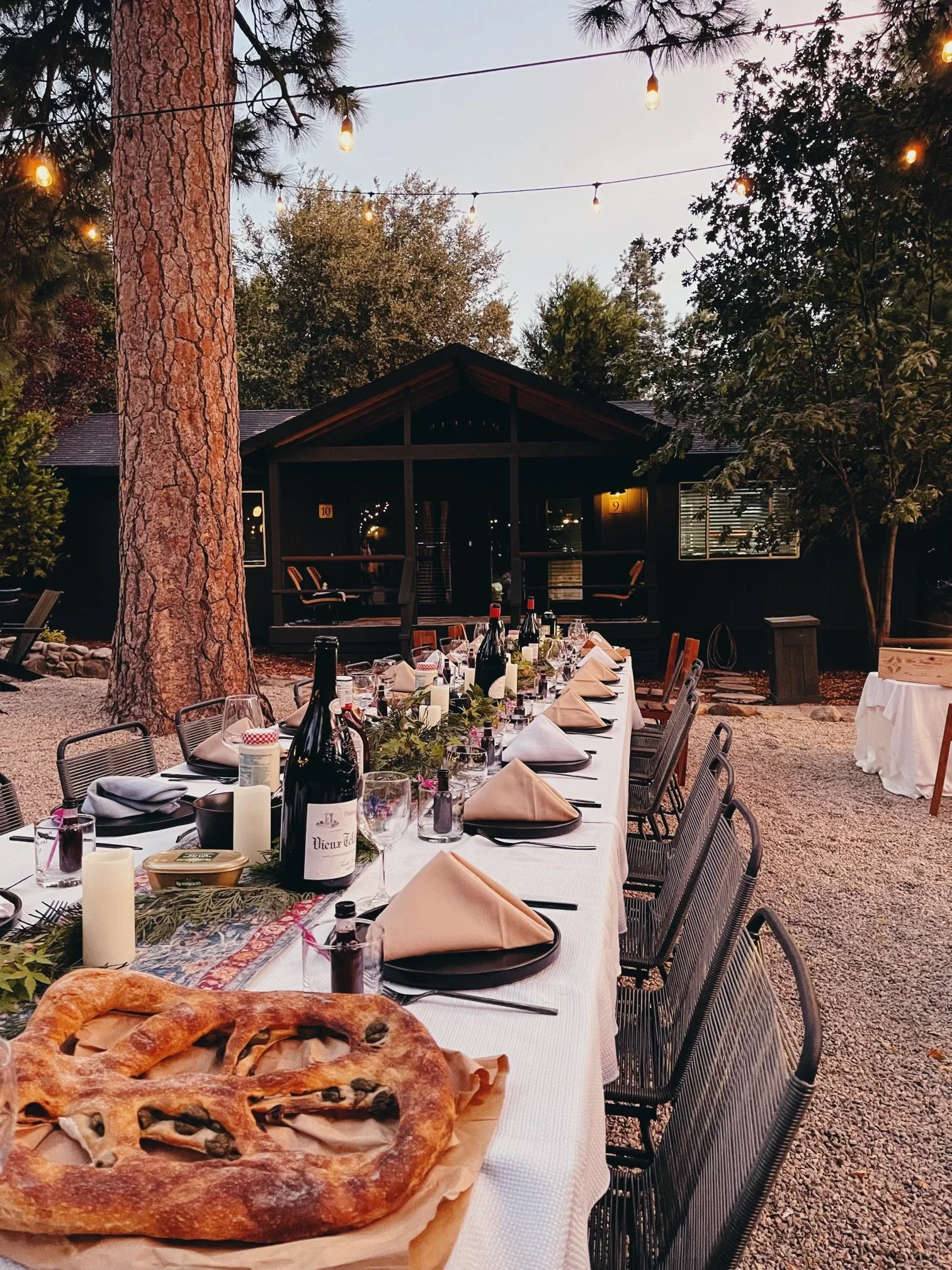 Long outdoor dining table set for a gathering, decorated with candles, wine bottles, glasses, and neatly folded beige napkins, situated in a wooded area with string lights overhead and a dark building in the background.
