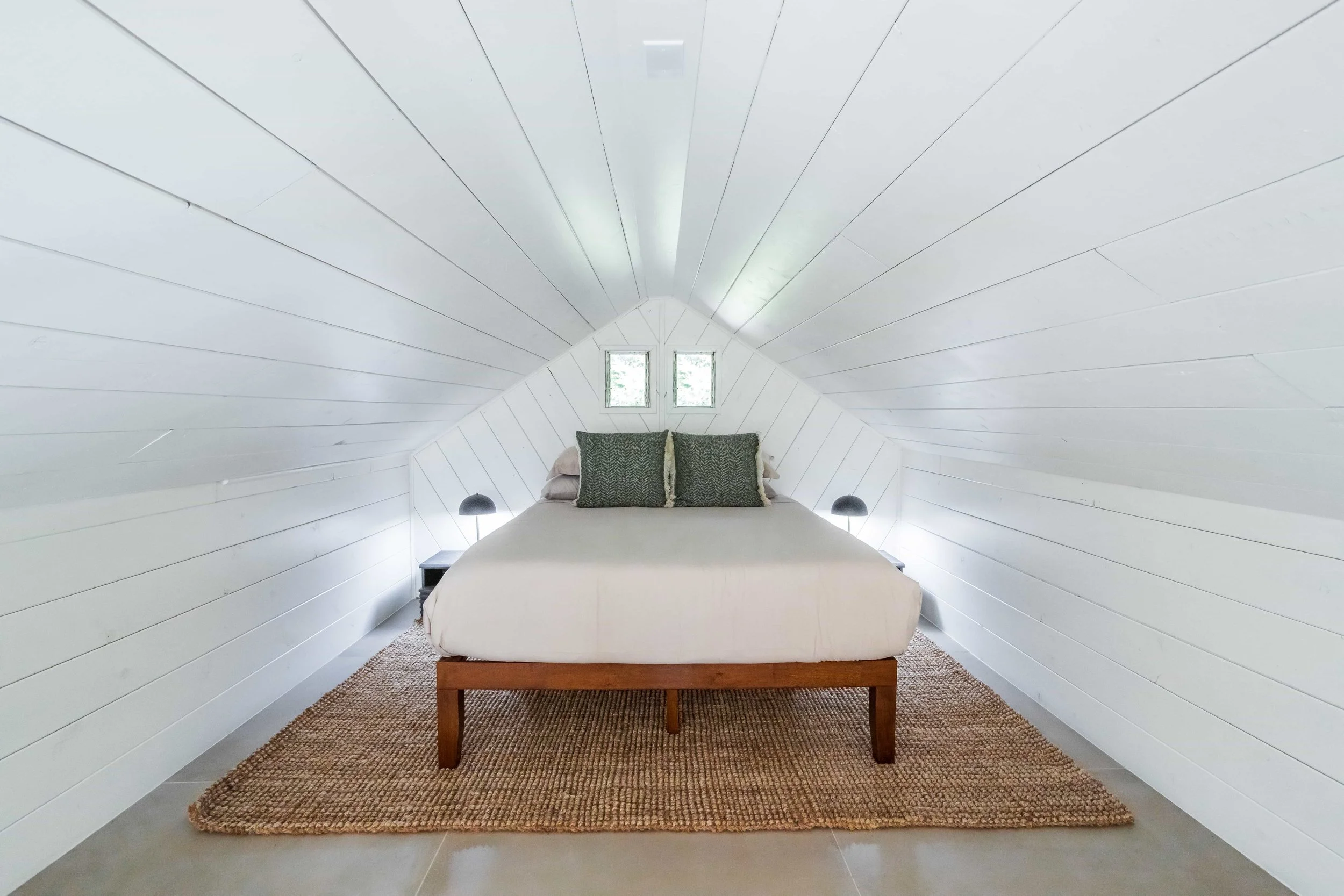 A minimalist bedroom in an attic with white painted wooden walls and ceiling, a bed with a white duvet and green pillows, small windows above the bed, side tables with black lamps, and a natural fiber rug on the floor.