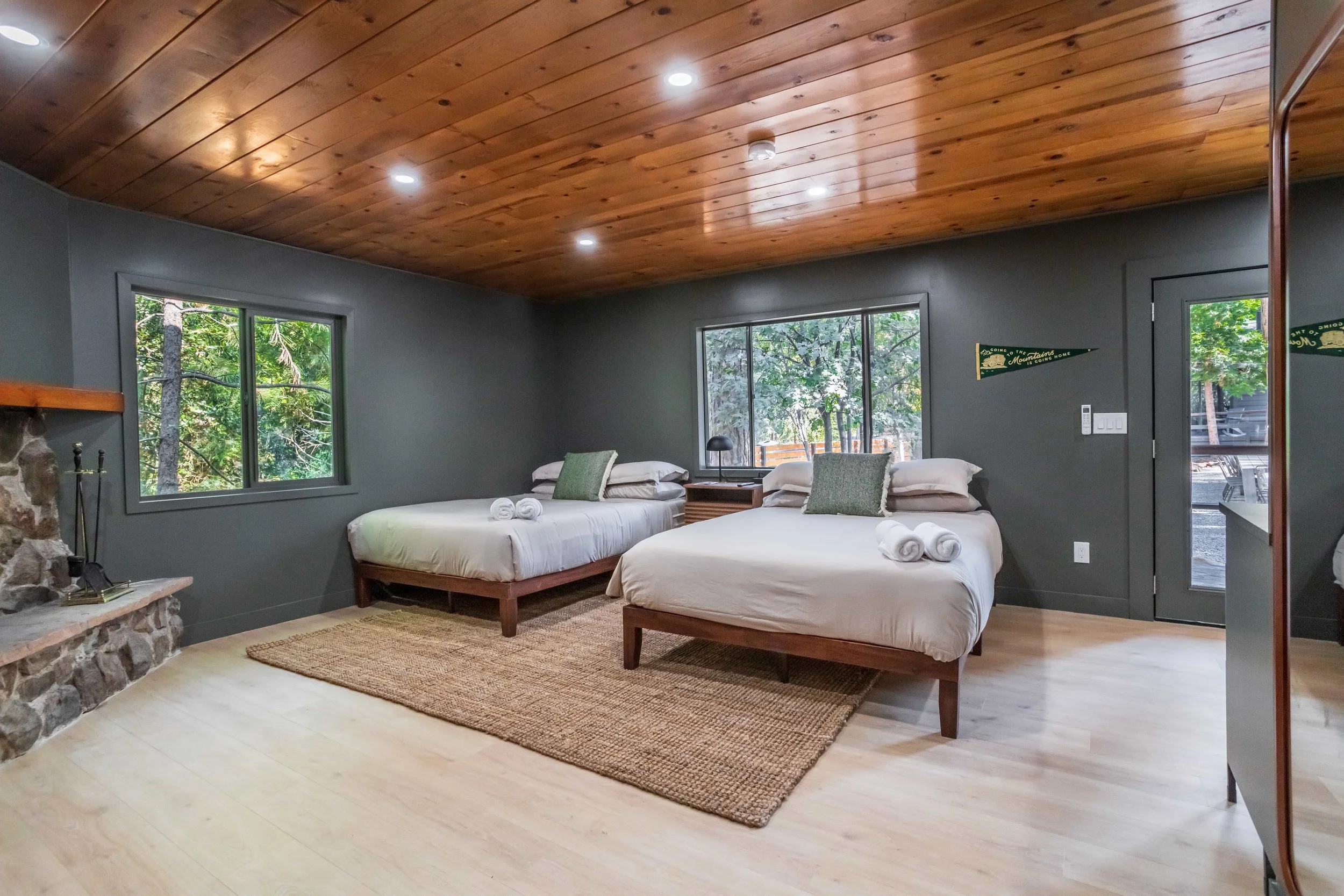 A bedroom with two beds, white bedding, green pillows, and rolled towels on each bed. There is a stone fireplace on the left, large windows showing trees outside, a wooden ceiling, and a beige rug on light-colored wood flooring.