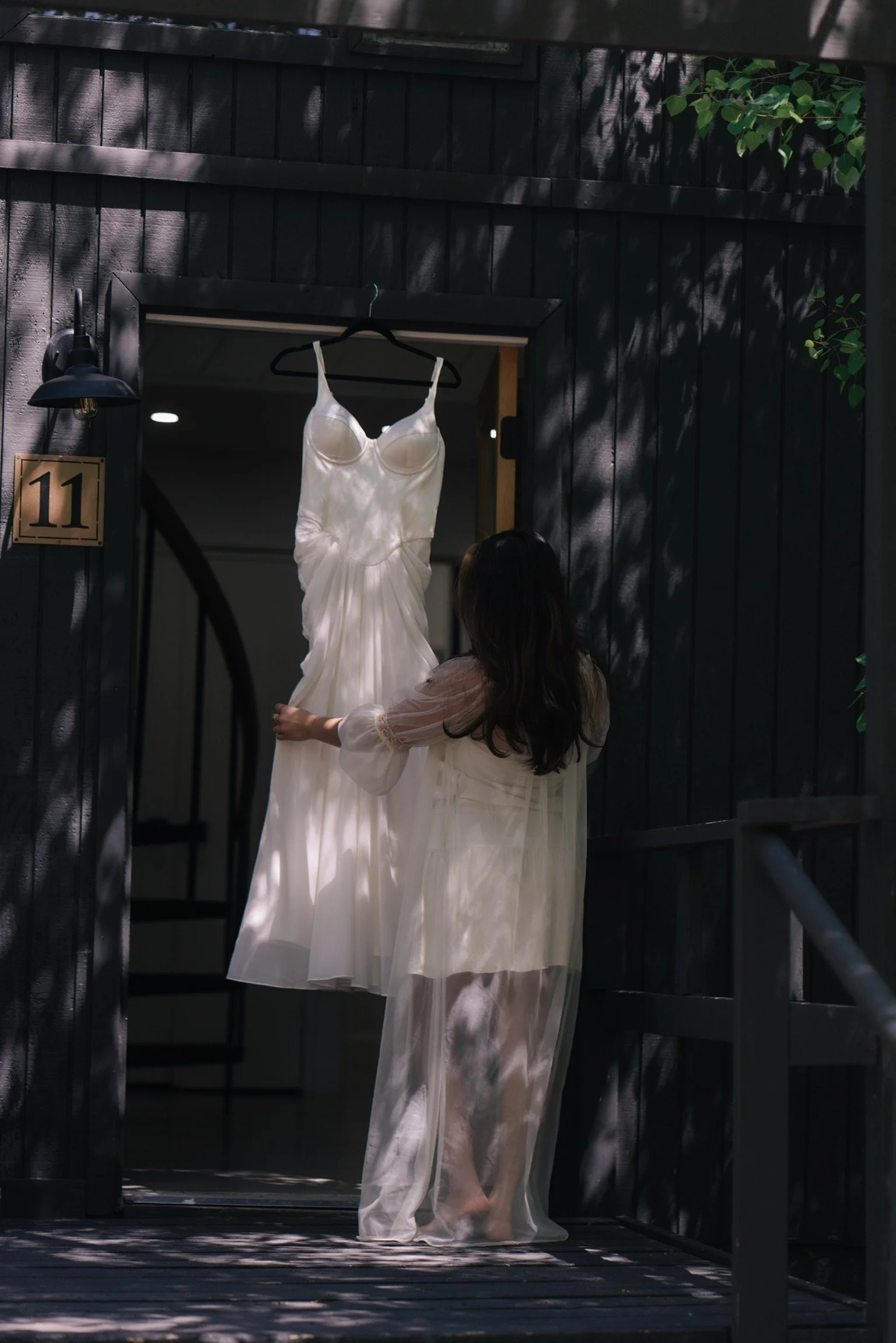 A woman in a sheer dress standing on a porch, holding a white dress hanging on a hanger.