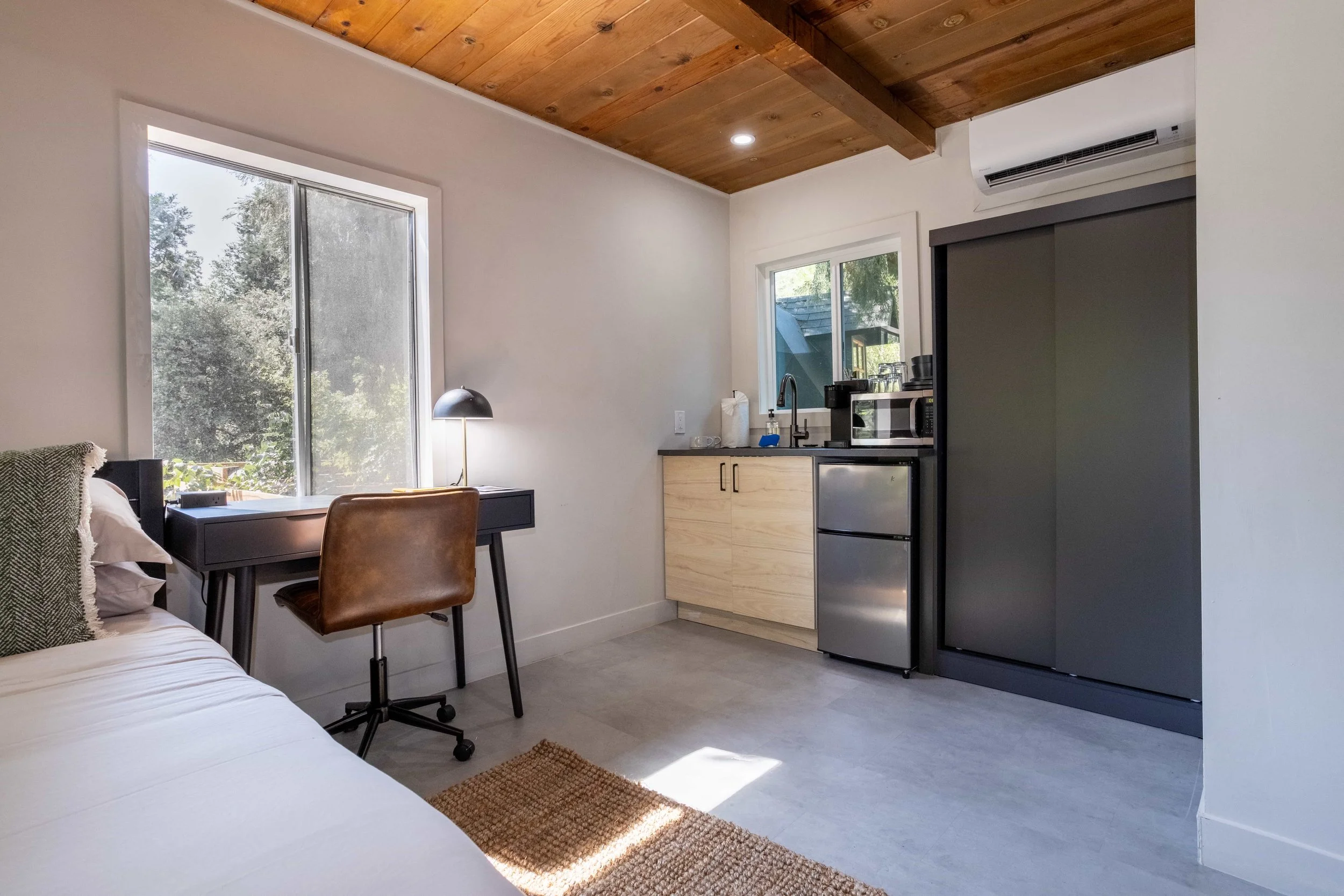 Small apartment studio with a bed, desk, chair, kitchenette, and large windows showing trees outside.