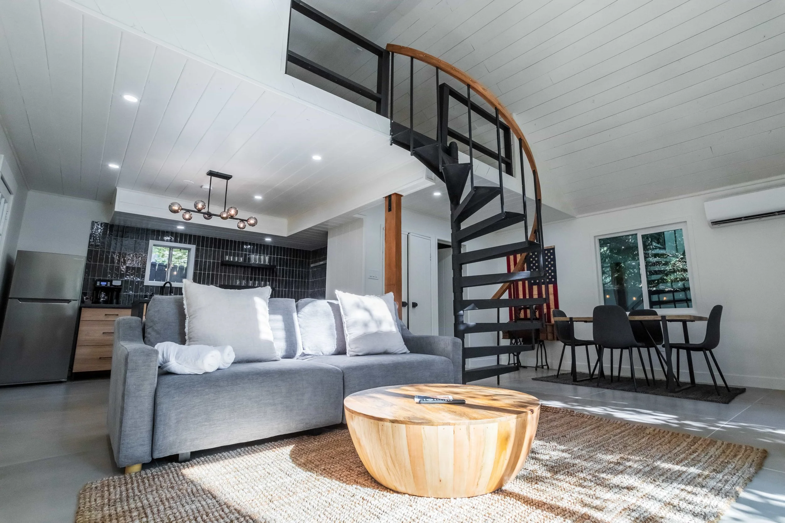Modern open-concept living room with a gray couch, wooden coffee table, and black spiral staircase leading to a second floor. Kitchen with black tiles and stainless steel refrigerator in the background. Dining area with a rectangular table and six bl