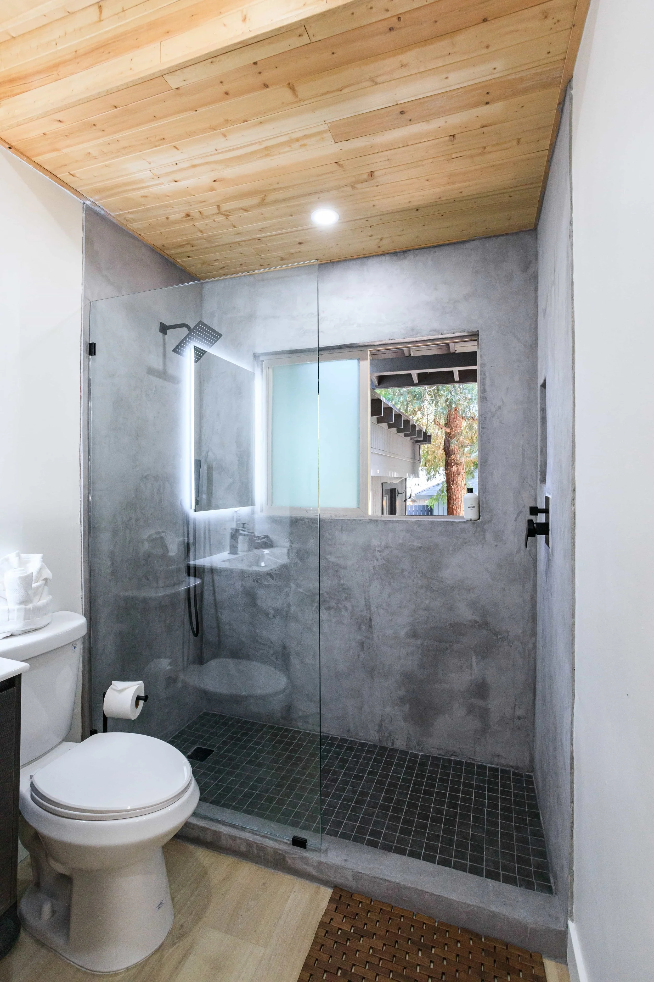 Modern bathroom with a walk-in shower, concrete walls, frosted window, wooden ceiling, and white toilet.