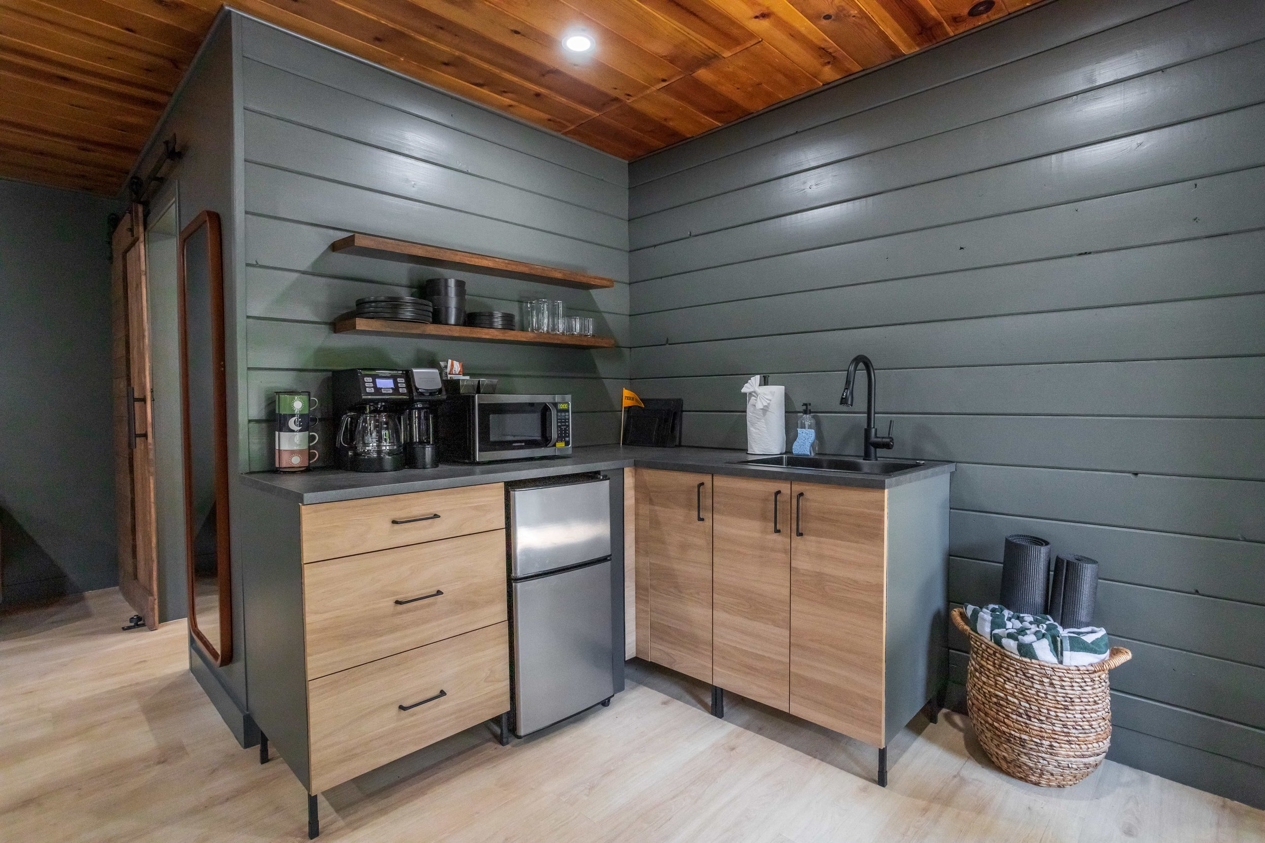 Mini kitchen with gray shiplap walls, wooden cabinetry, black countertop, small refrigerator, coffee maker, microwave, shelves with dishes, and a basket of rolled mats or towels.