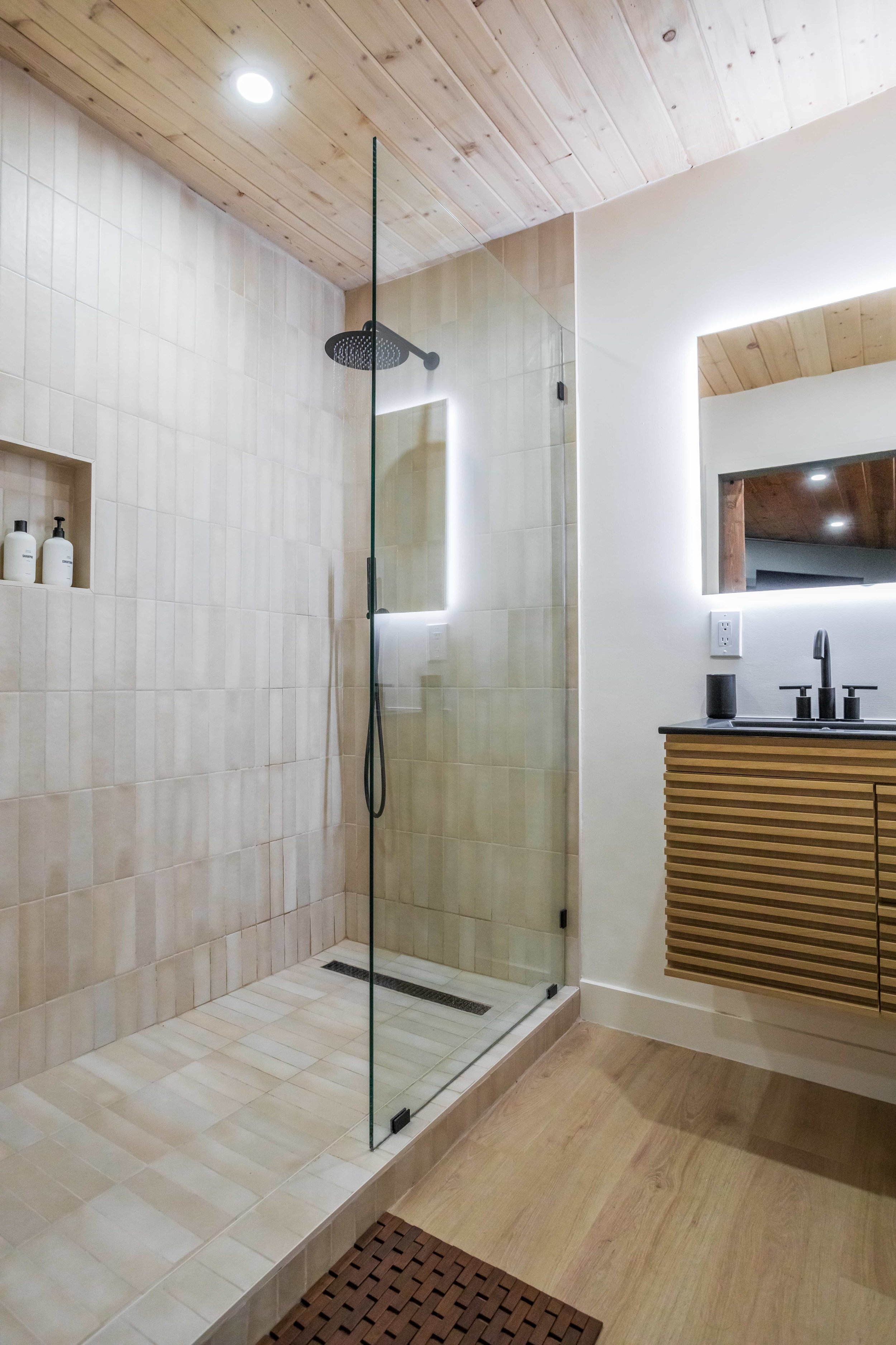 Modern walk-in shower with beige tiles, glass door, rain showerhead, wooden ceiling, and a vanity with a mirror.