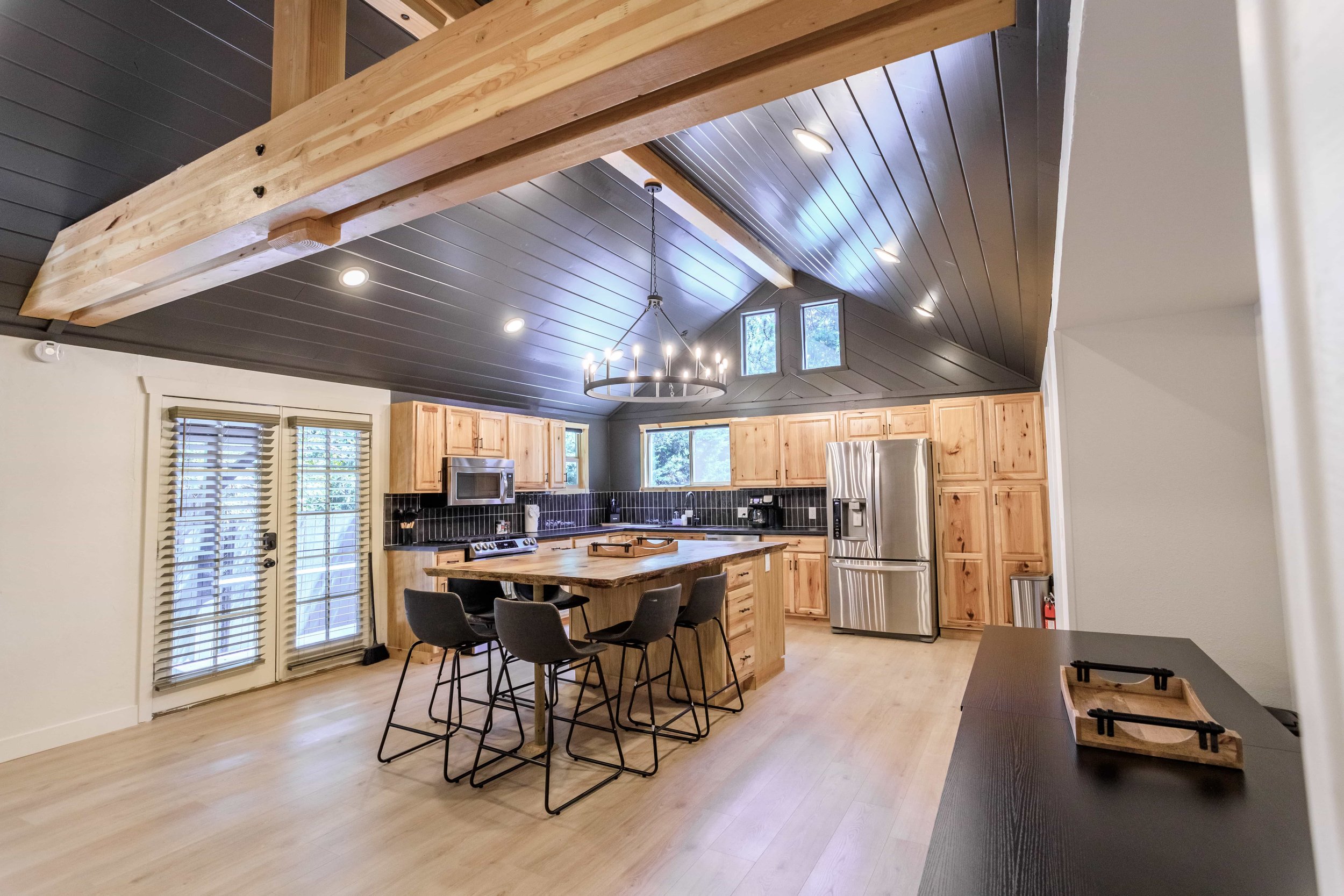 A spacious kitchen with wooden cabinets, black backsplash, stainless steel appliances, black chairs around a central island, and a black ceiling with recessed lighting and a chandelier.