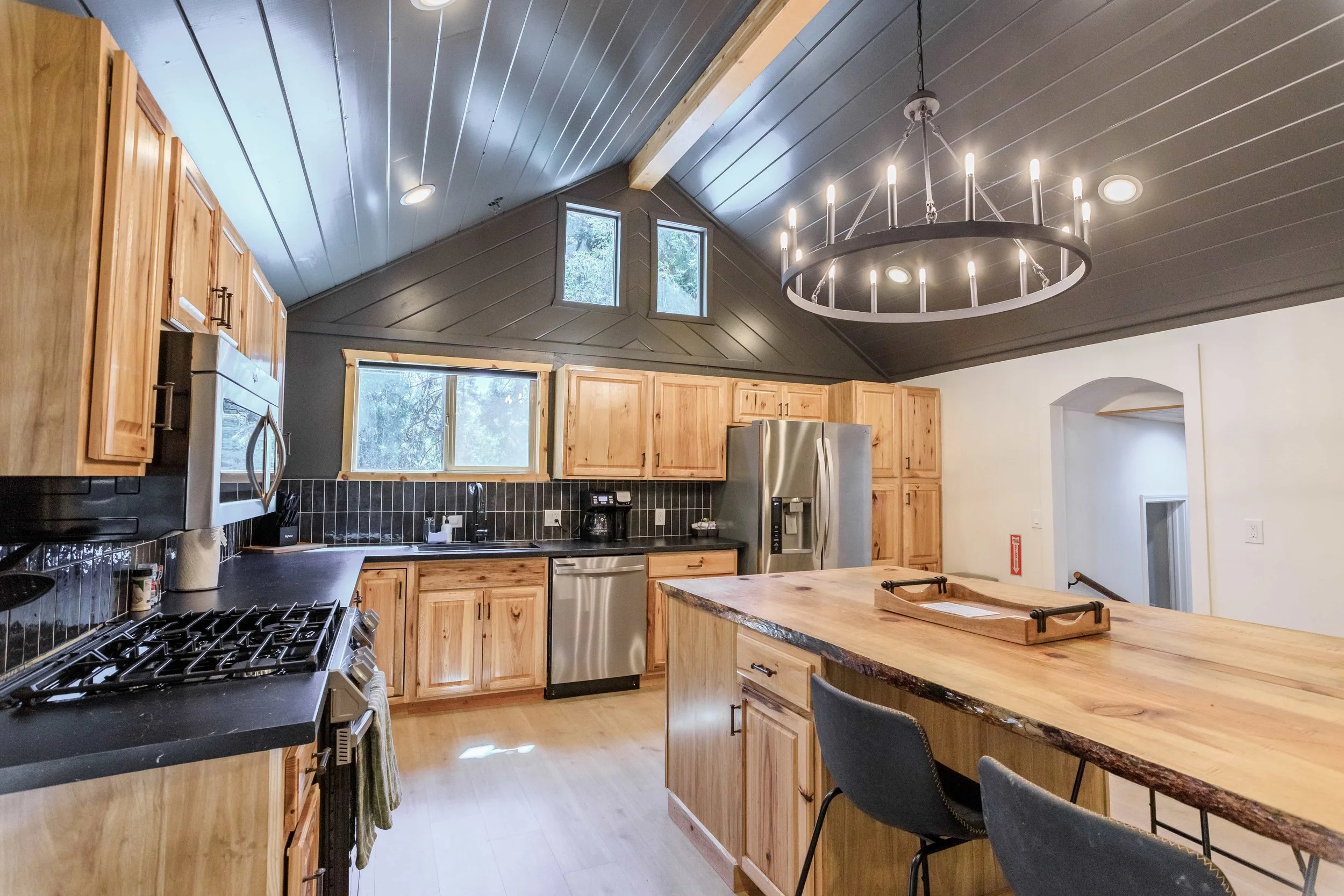 Kitchen with wooden cabinets, black countertops, stainless steel appliances, black tiled backsplash, large wooden island, black chairs, gray ceiling, modern chandelier, windows with natural light.