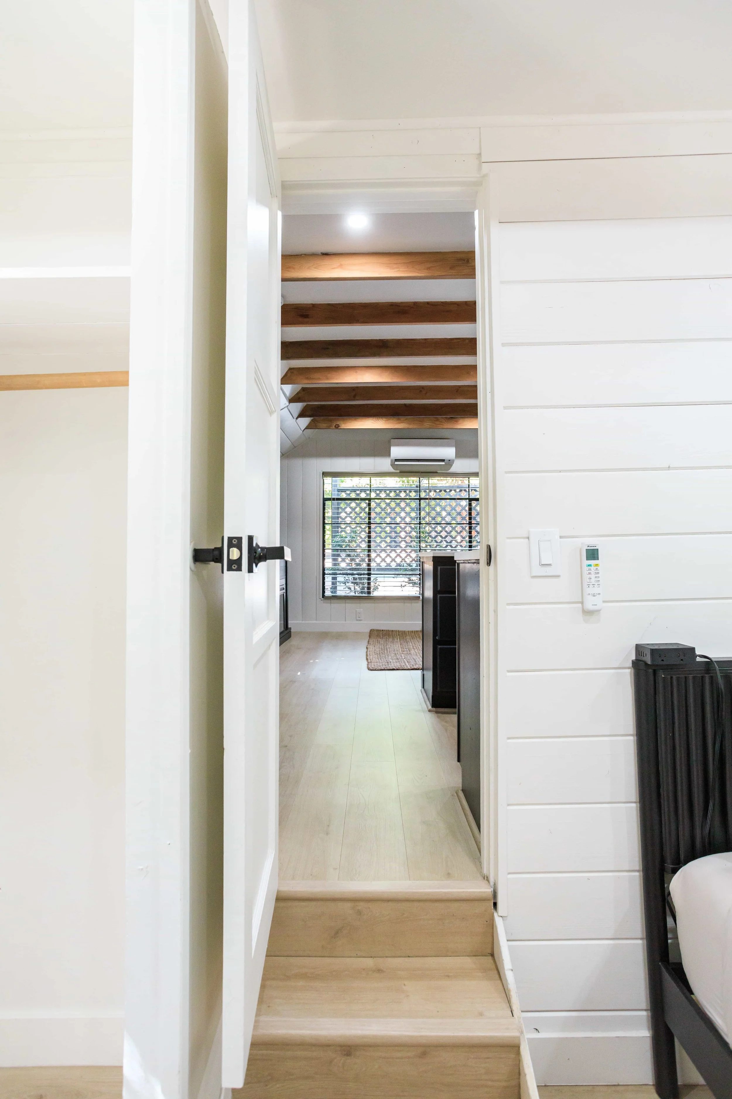 View from doorway into a bright room with wooden ceiling beams, window with black security bars, and a tan rug on light wood flooring.
