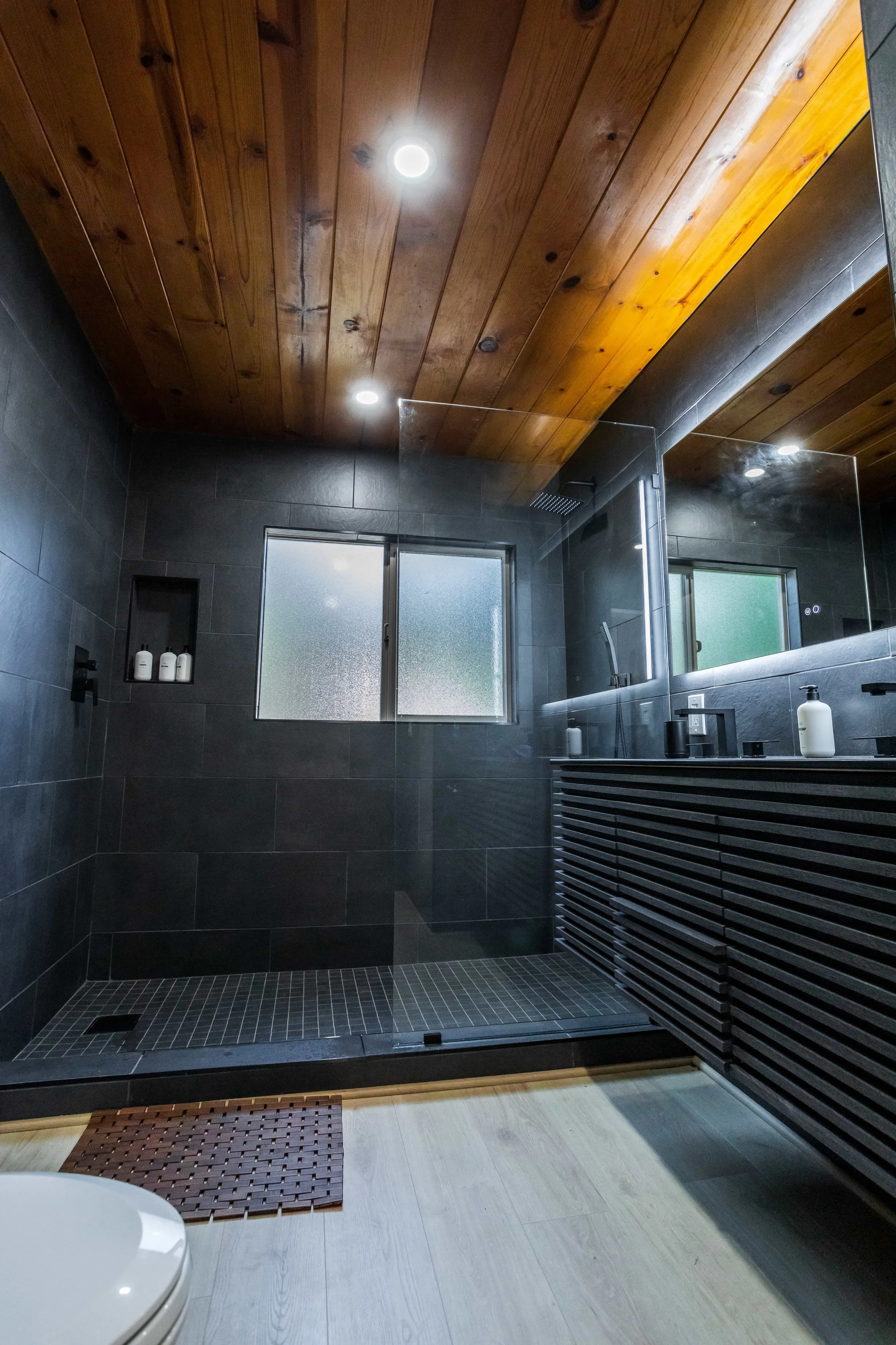 The Pinetree Hotel Idyllwild:  a - Idyllwild California, cozy cabin, mountain resort, mountain getaway Modern bathroom in cabin 1 with dark tiled walls, wooden ceiling, and walk-in shower with glass door and frosted window.