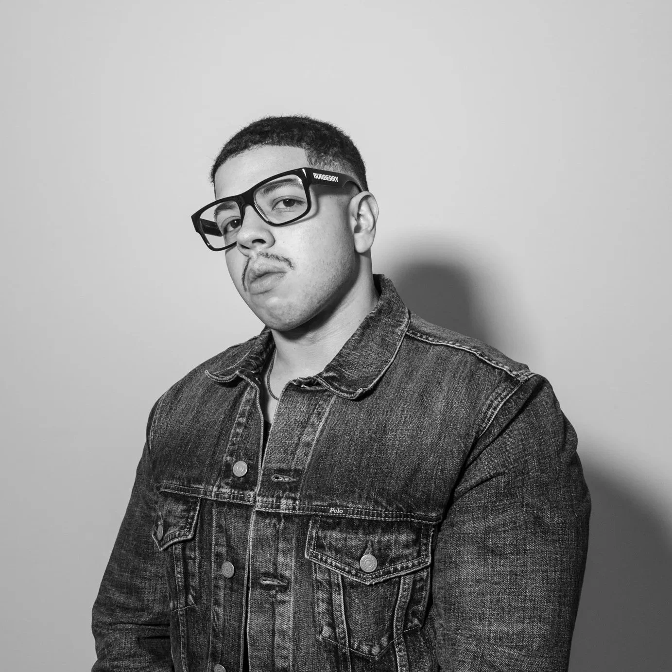 Black and white portrait of a young man with short hair, wearing glasses and a denim jacket, looking slightly to the side.
