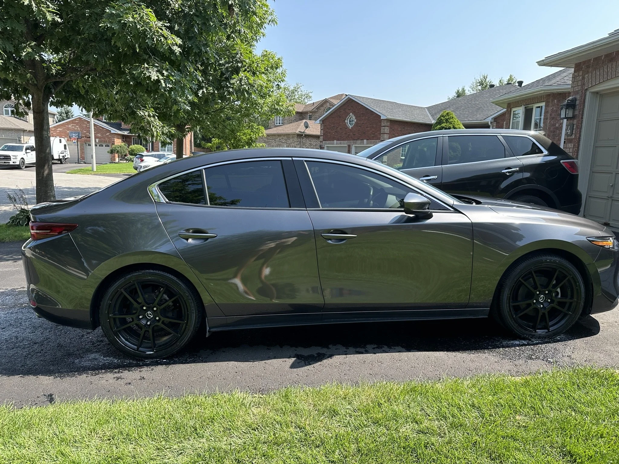 A gray sedan parked on a residential driveway, with a black SUV parked behind it. There are houses, trees, and a clear blue sky in the background.