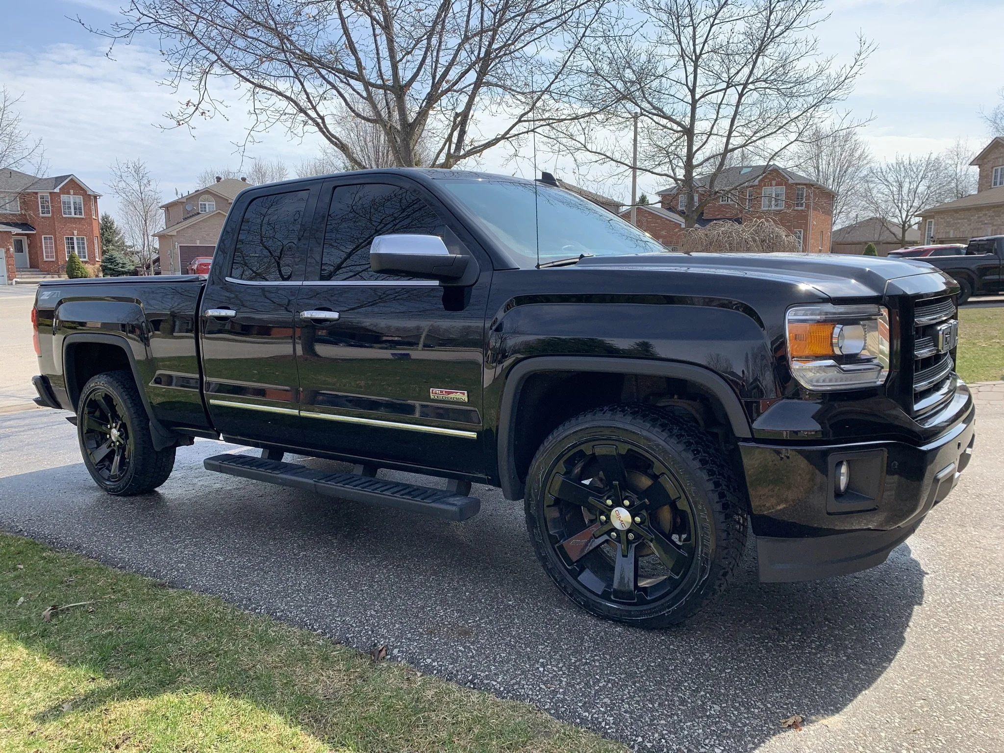A black pickup truck parked on a driveway with a suburban neighborhood in the background, including houses, trees, and a clear sky.