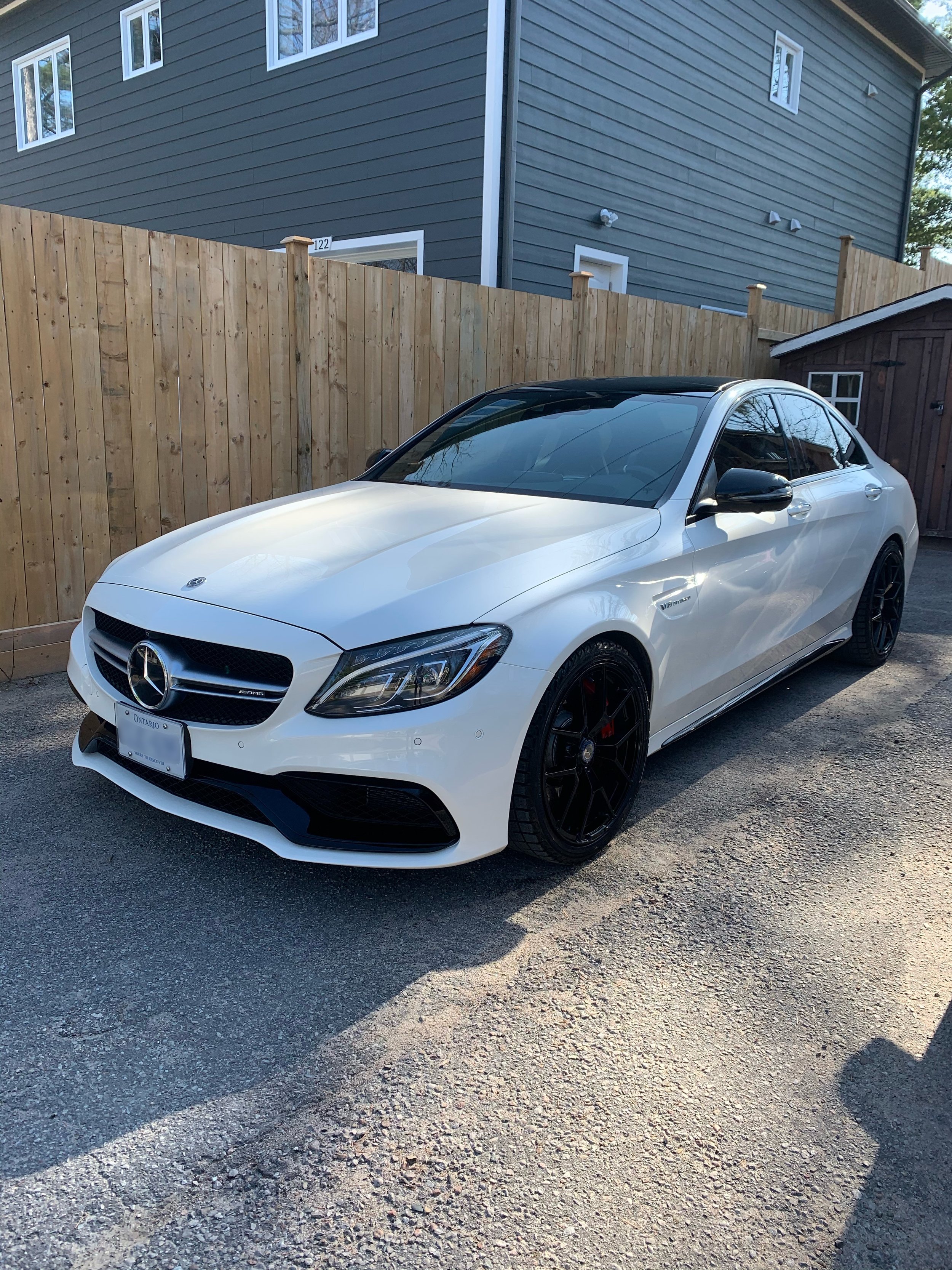 White Mercedes-Benz coupe parked on a gravel driveway beside a wooden fence and a blue house, with a small wooden shed nearby.