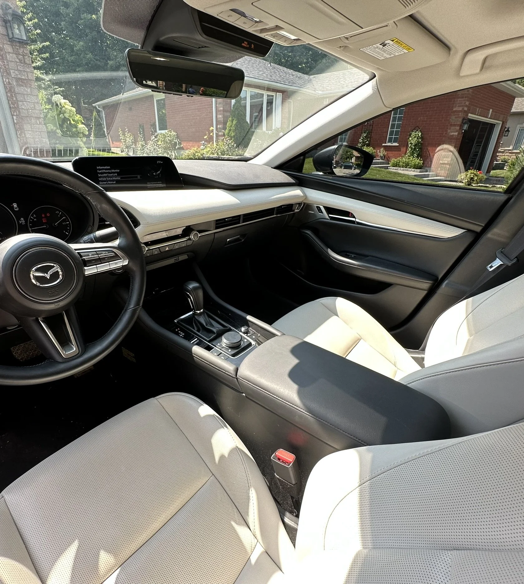 Interior of a modern Mazda vehicle showing the dashboard, steering wheel, gear shift, and front seats with light-colored upholstery. Outside view of residential houses through the windshield.