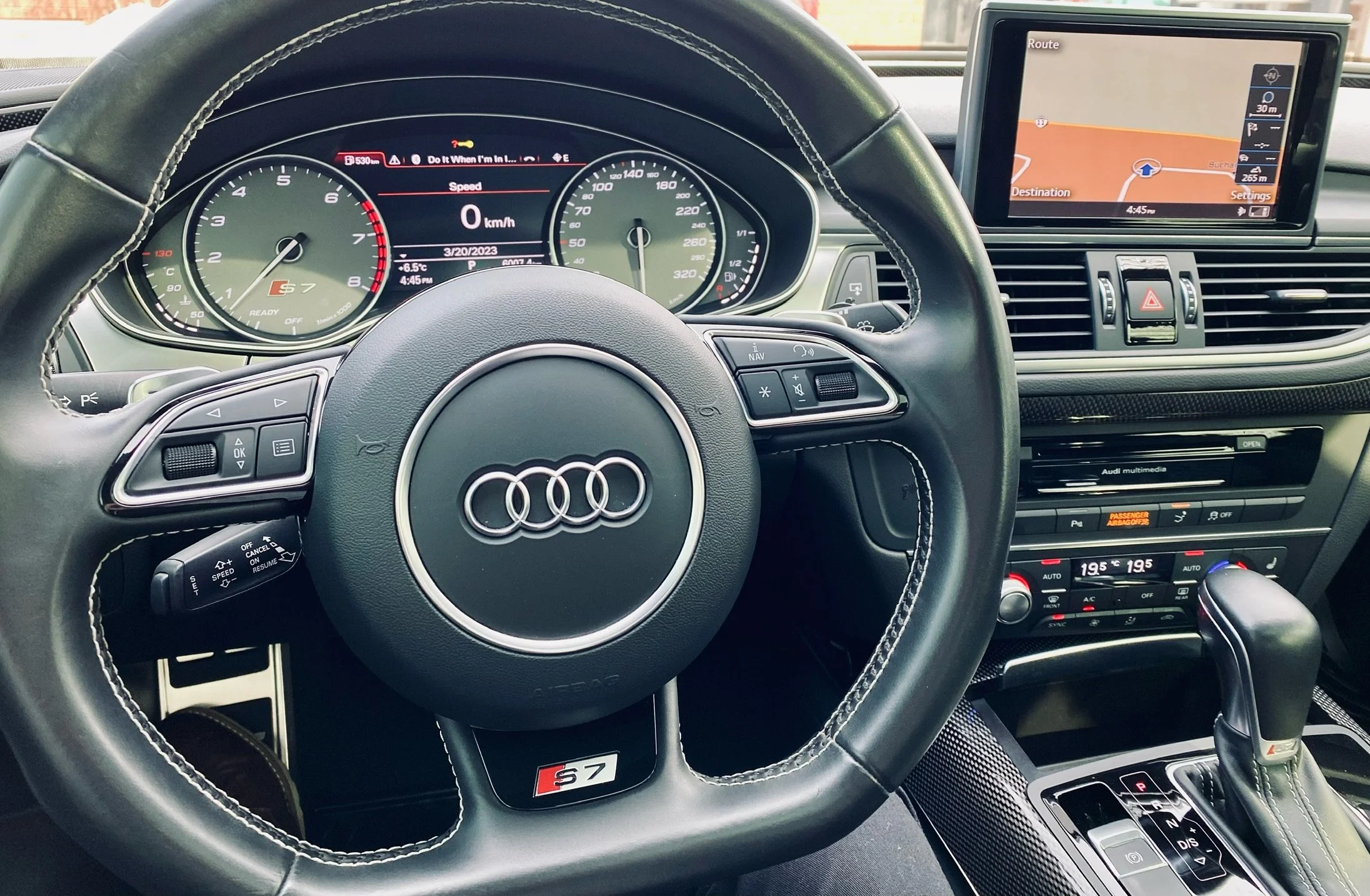 The interior of an Audi S7, showing the steering wheel, dashboard, and navigation screen. The steering wheel has Audi's logo and 'S7' badge. The dashboard displays a speed of 0 km/h, with a digital tachometer and other gauges. The navigation screen s