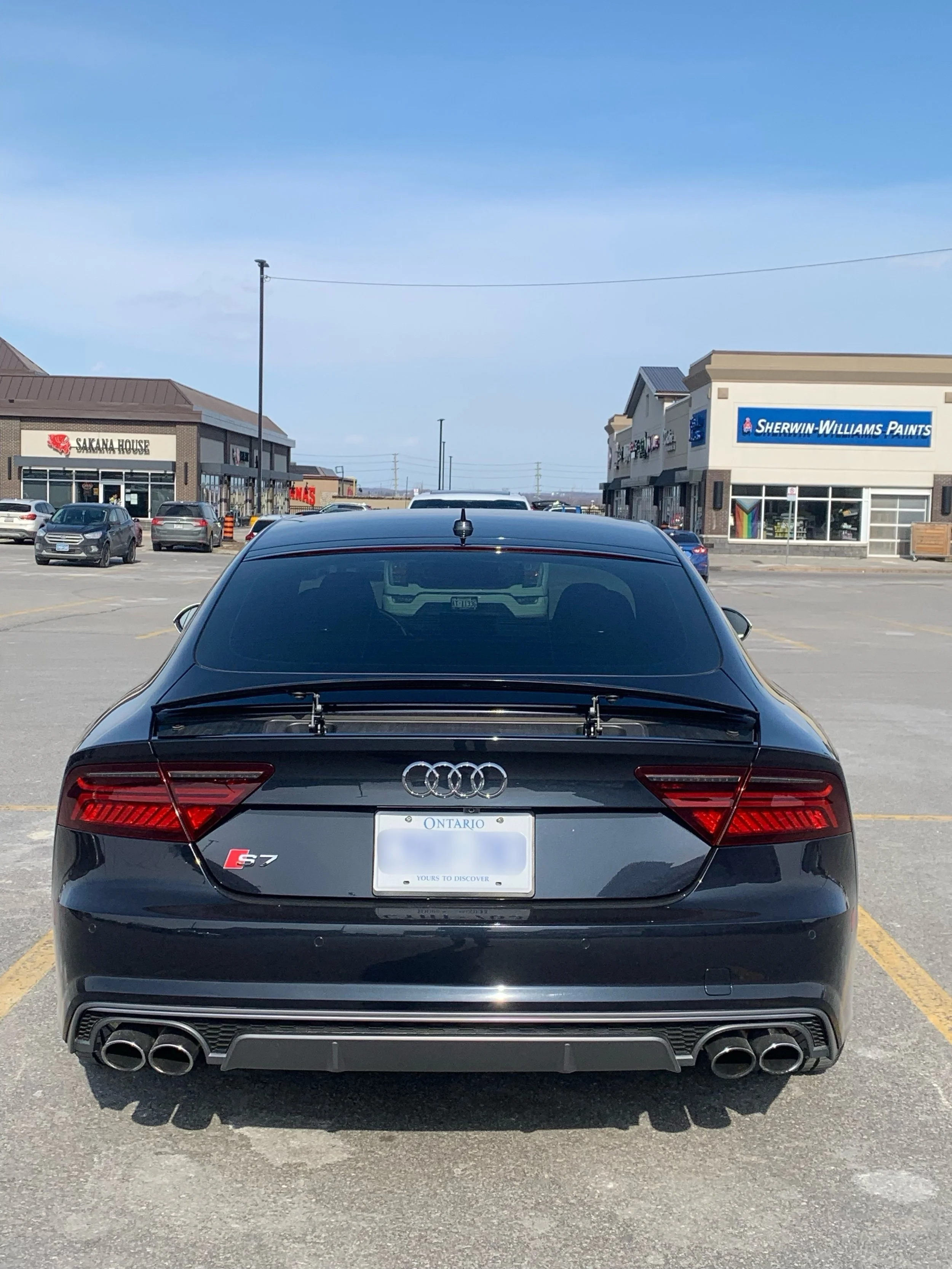 Black Audi S7 sedan parked in a shopping center parking lot with stores like Sherwin-Williams Paints and Sakana House visible in the background.