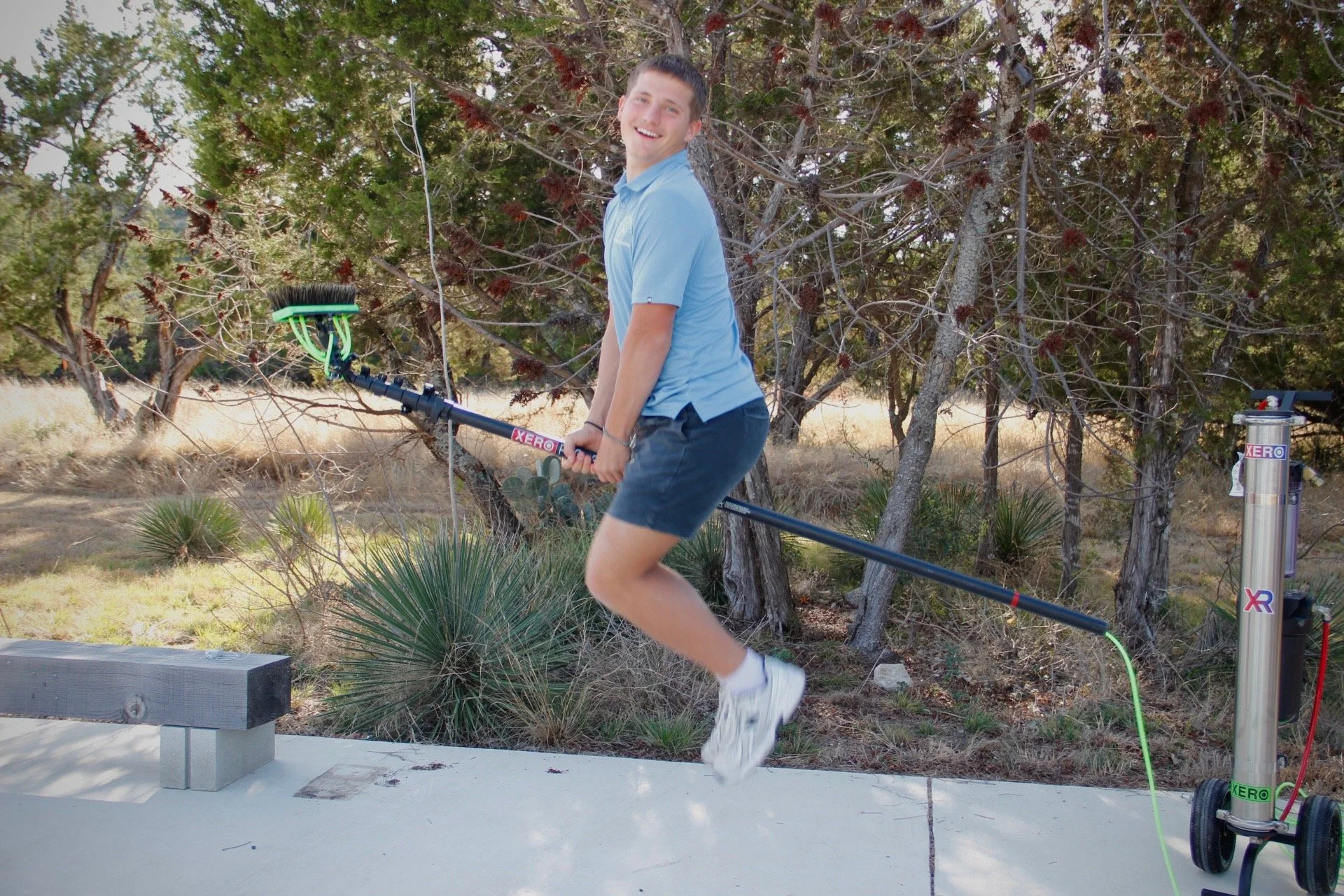 A young man with short hair, wearing a light blue polo shirt, dark shorts, and white sneakers, is jumping with a metal detector in an outdoor wooded area.