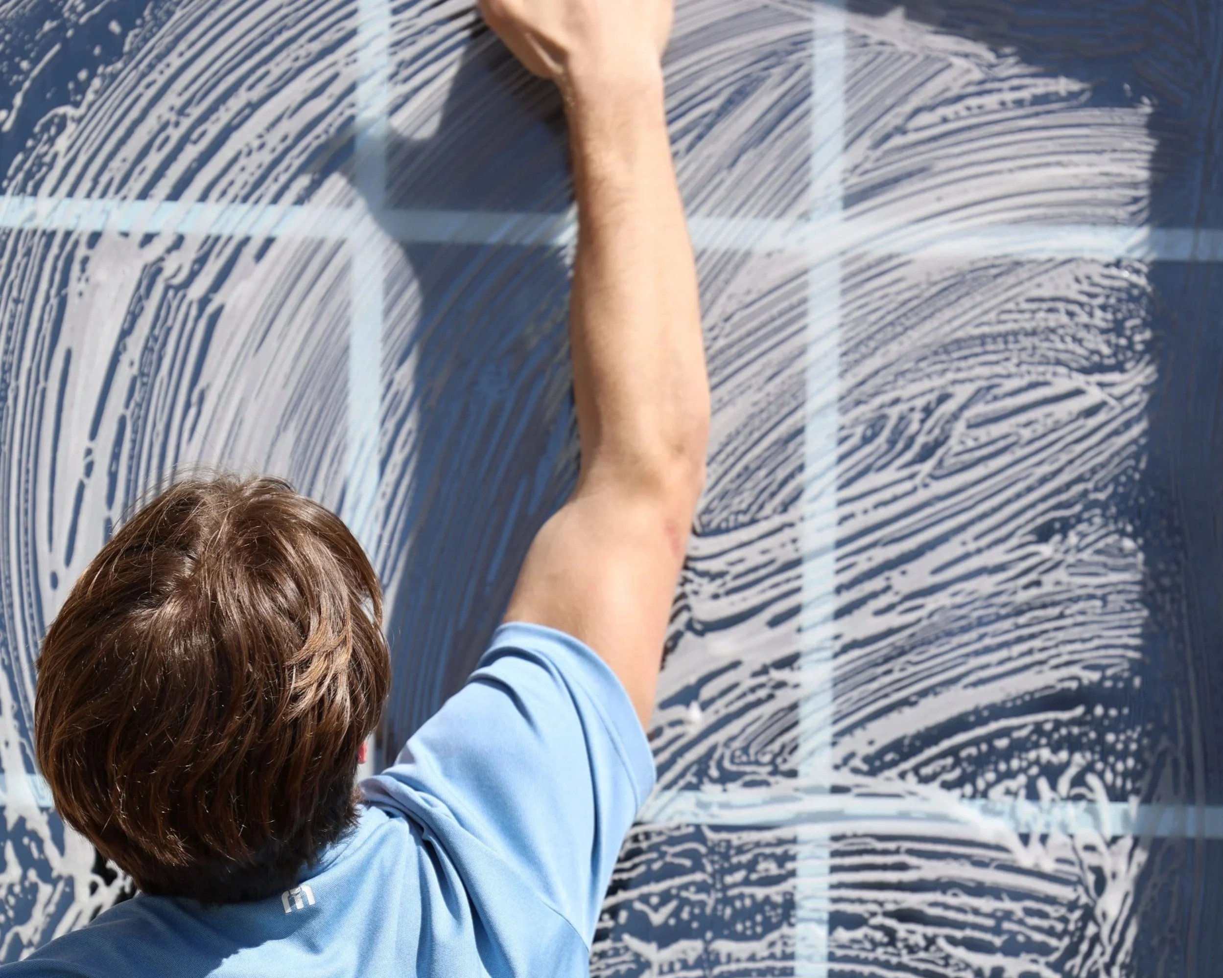 A person with brown hair in a blue shirt is cleaning a large window with soap