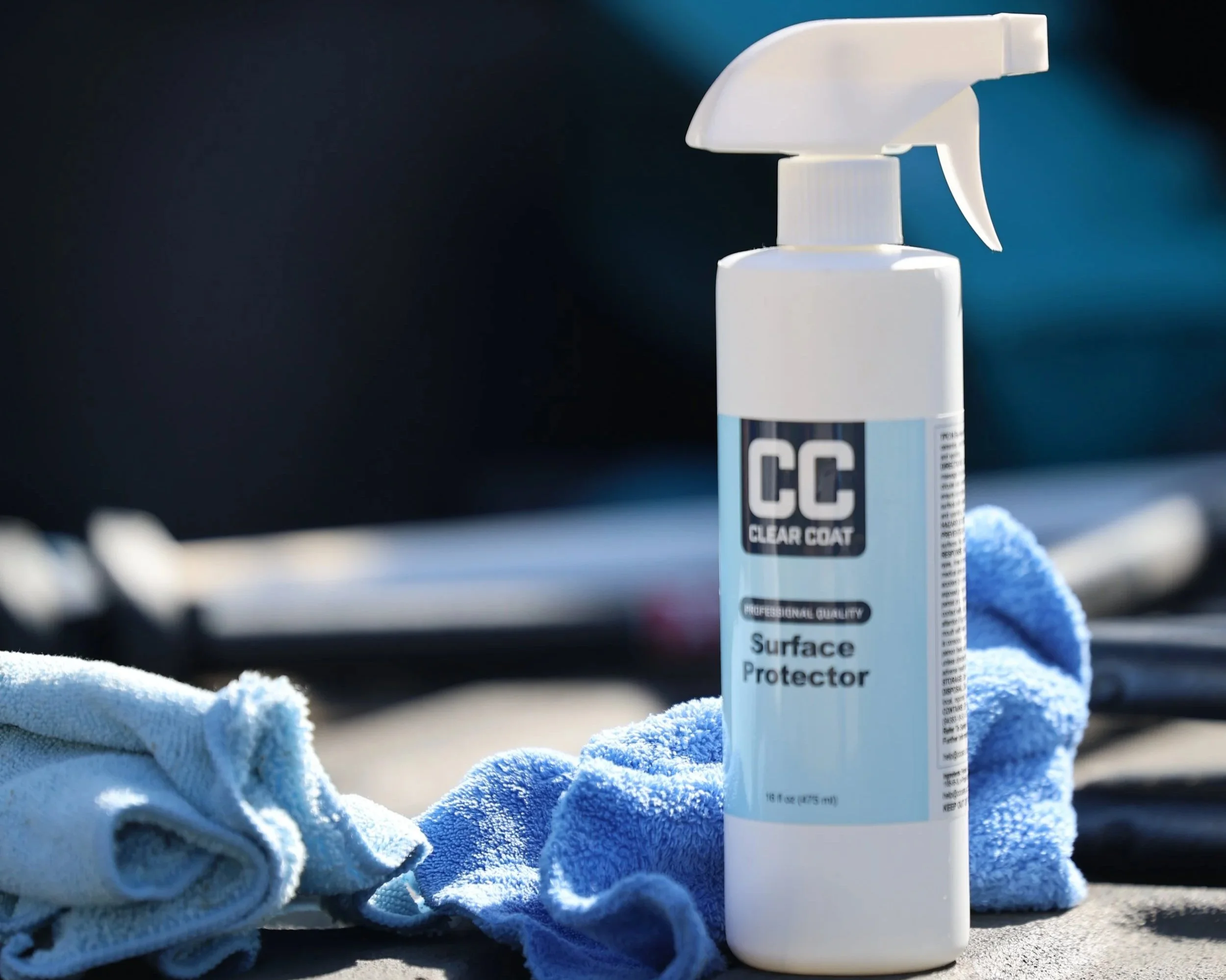 A spray bottle labeled 'CC Clear Coat Surface Protector' with a blue towel and a white cloth on a gray surface.