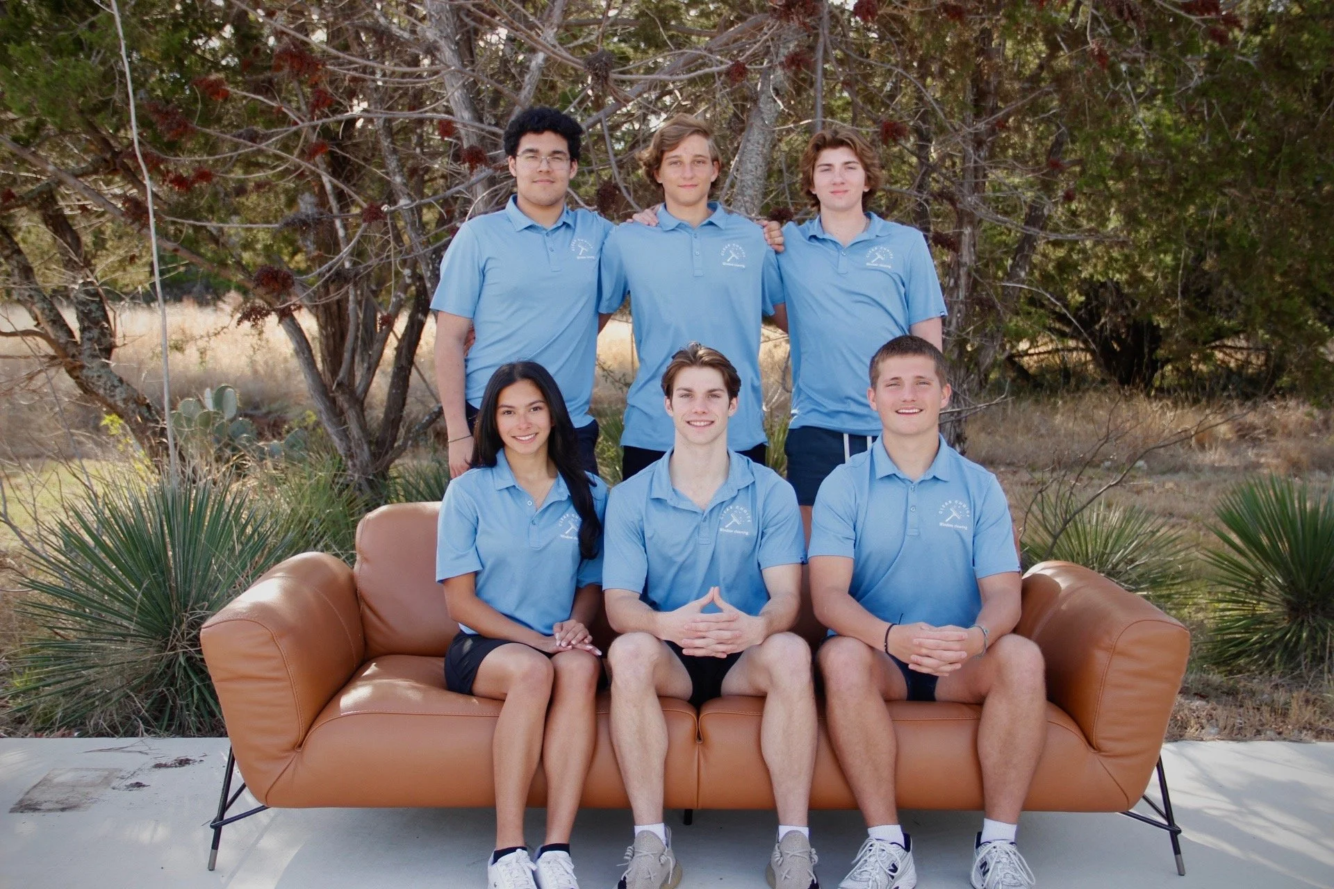 Group of six teenagers wearing matching blue polo shirts, sitting and standing outdoors with trees and desert plants background.