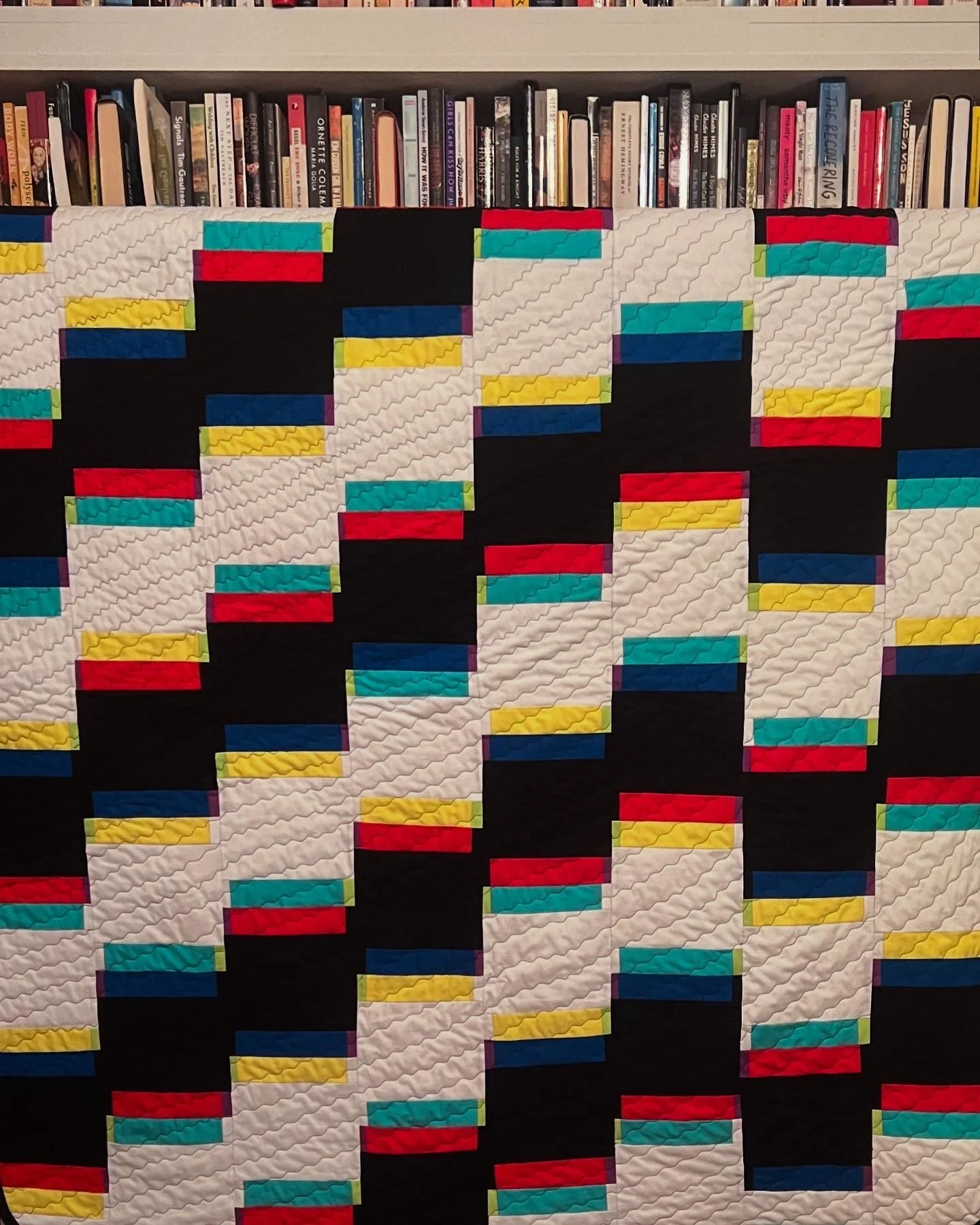 A colorful quilt with a geometric pattern of rectangles in red, yellow, blue, teal, and purple on a black background, hanging in front of a bookshelf filled with books.