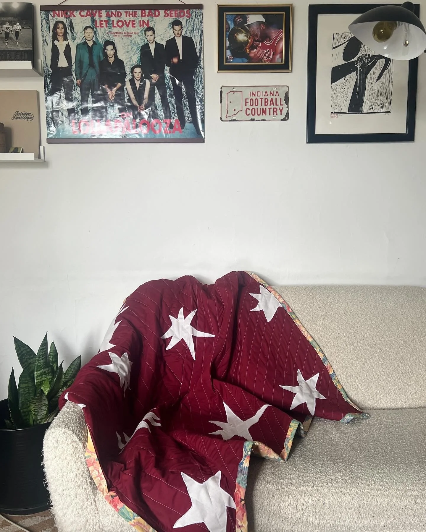 A beige couch with a red quilt featuring white stars draped over it, next to a black potted plant, against a white wall decorated with music posters, a framed sports photo, and a black and white artwork. A standing lamp with a black shade and a white