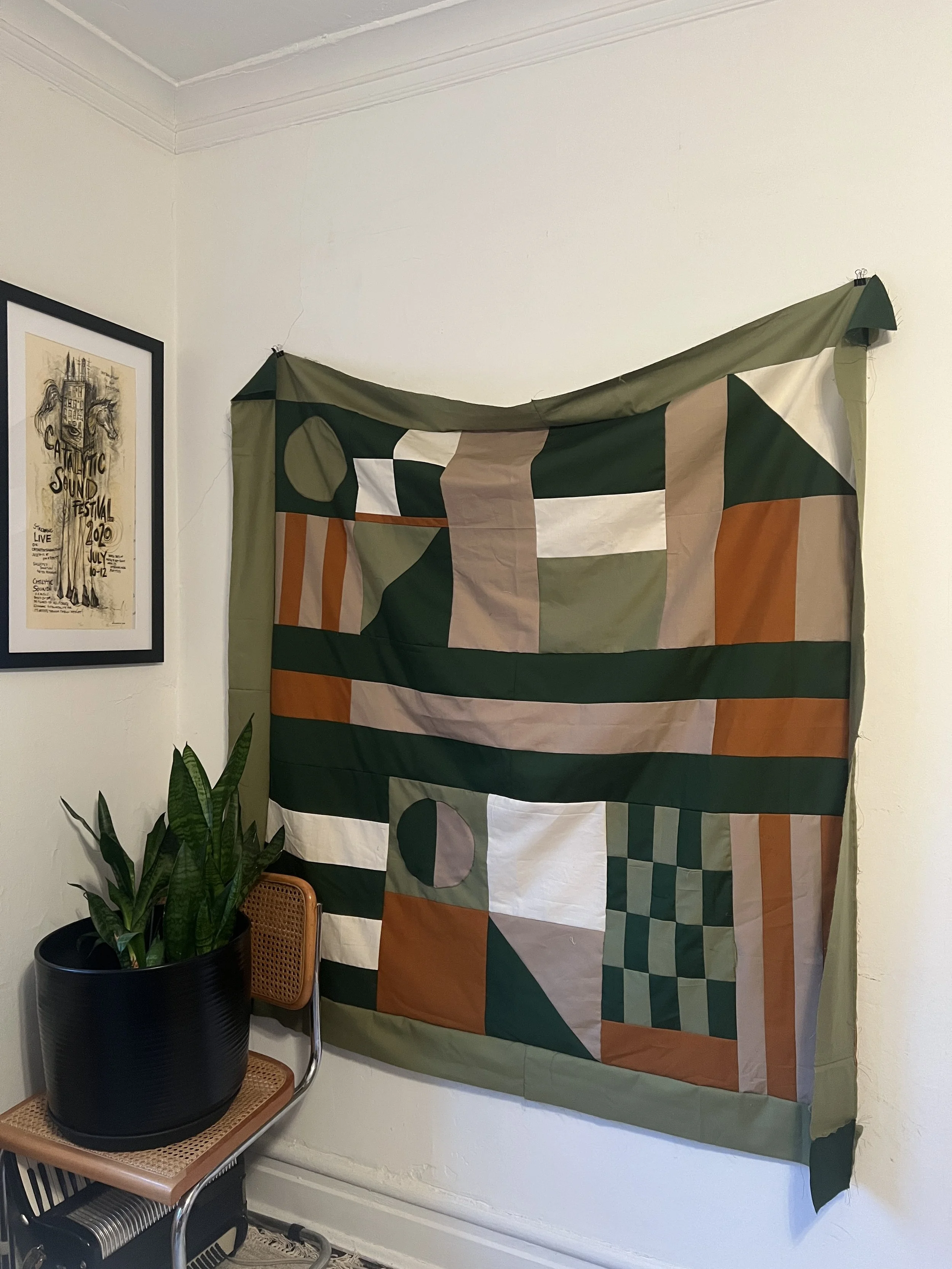 A colorful abstract quilt hanging on a white wall in a room. There is a framed poster on the left side and a potted snake plant on a chair below the quilt.
