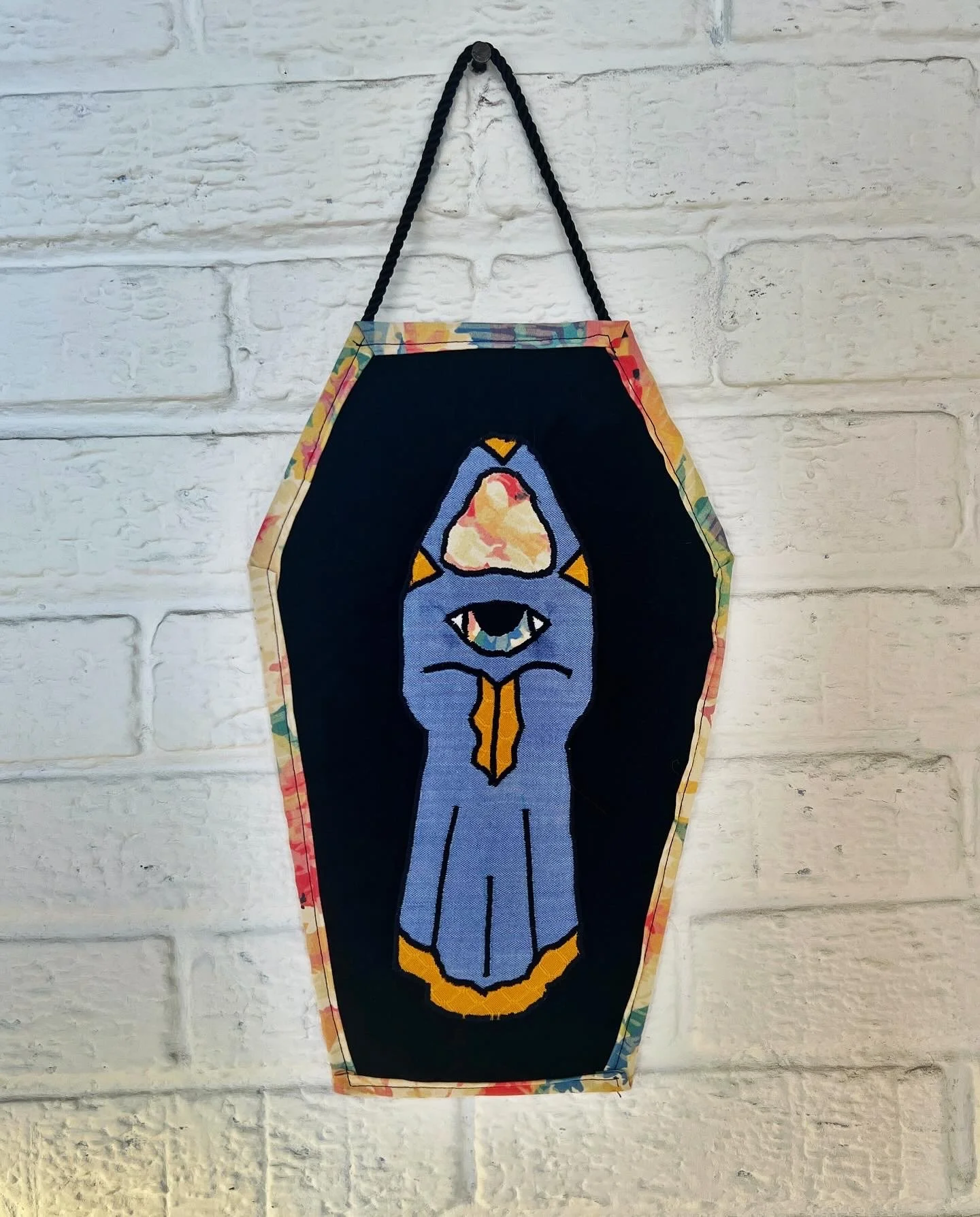 Decorative hanging textile art piece featuring a stylized cat face with closed eyes and a prominent third eye on the forehead, bordered with multi-colored floral fabric, hanging on a white brick wall.