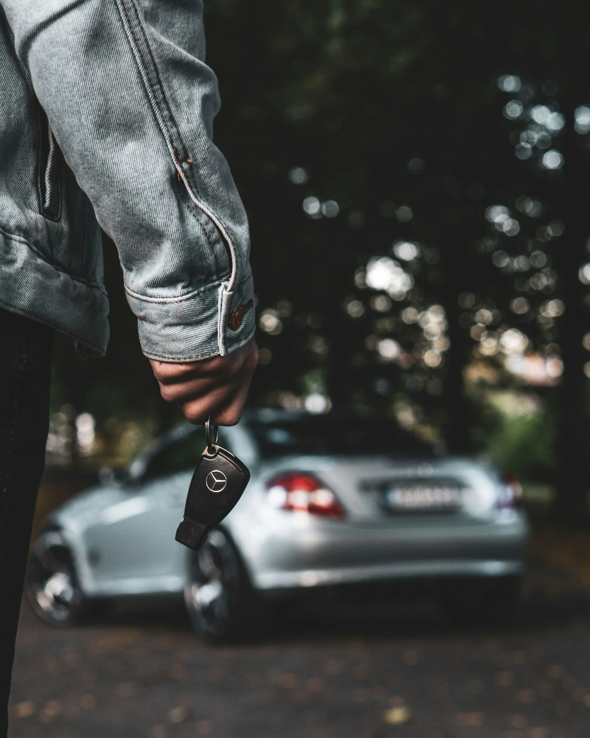 Close-up of a person's arm in a gray denim jacket holding a Mercedes-Benz car key, with a silver Mercedes-Benz car in the background, slightly out of focus, parked outdoors among trees.