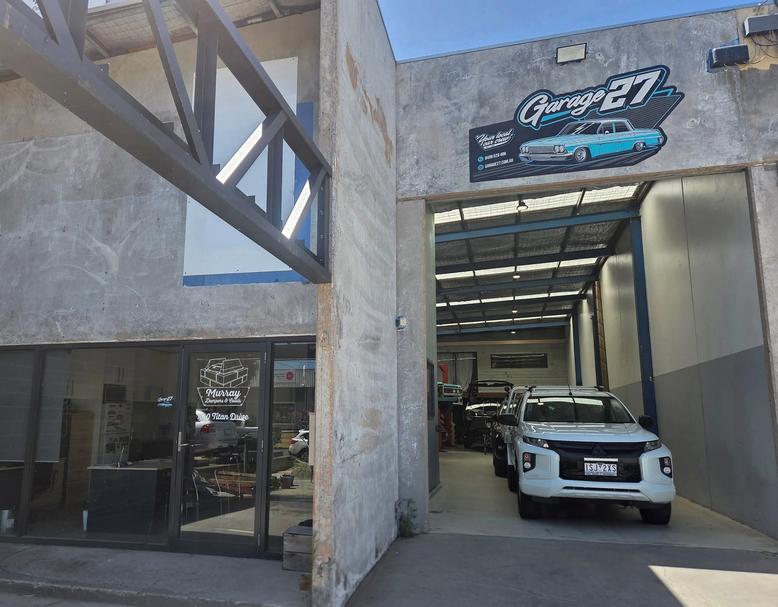 Exterior of an auto repair shop with a sign reading 'Garage 27' and a classic blue car painted on the sign. The shop has open garage bays with vehicles inside, including a white SUV with a license plate, and a glass entrance door.