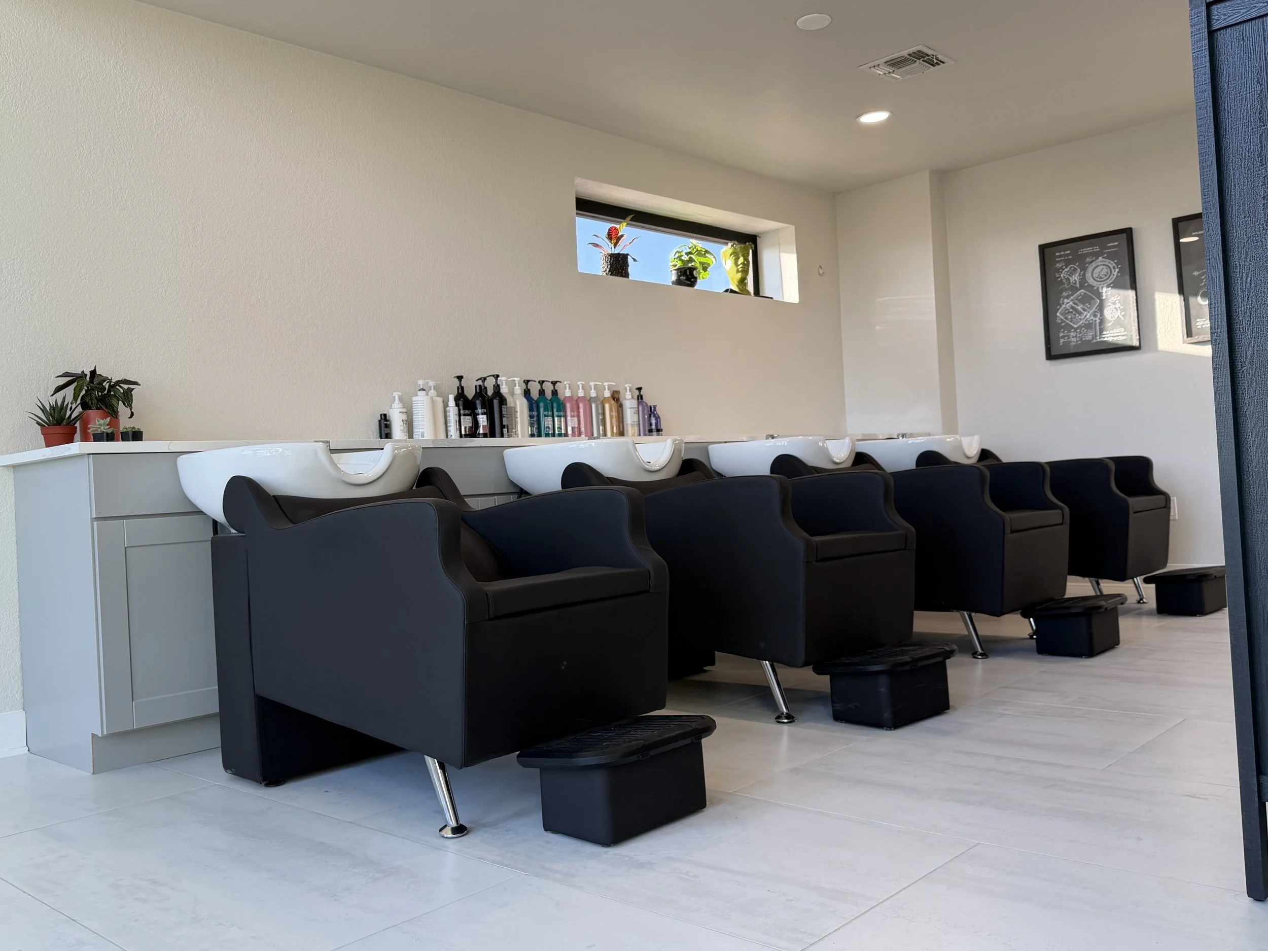 Interior view of a hair salon with three black salon chairs in front of white washbasins, bottles of hair products on a white counter, and a small window with decorative plants.