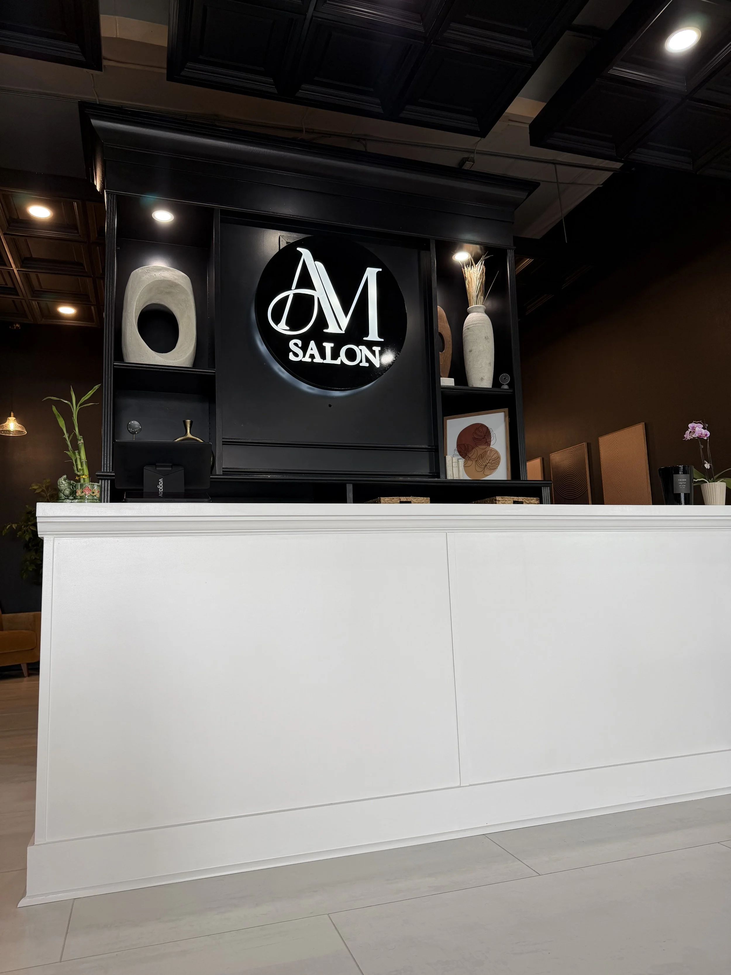 Reception area of a salon with white counter, black wall decor, and a round sign with "AM SALON" in illuminated letters, decorated with vases and abstract art.