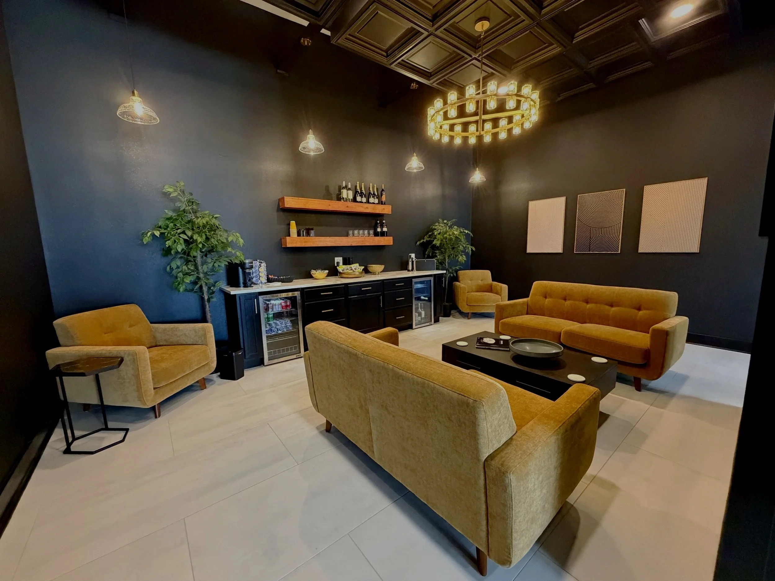 A modern lounge room with black walls, yellow velvet couches, and a round chandelier. There are two potted plants, abstract wall art, and a beverage bar with shelving and mini refrigerator.