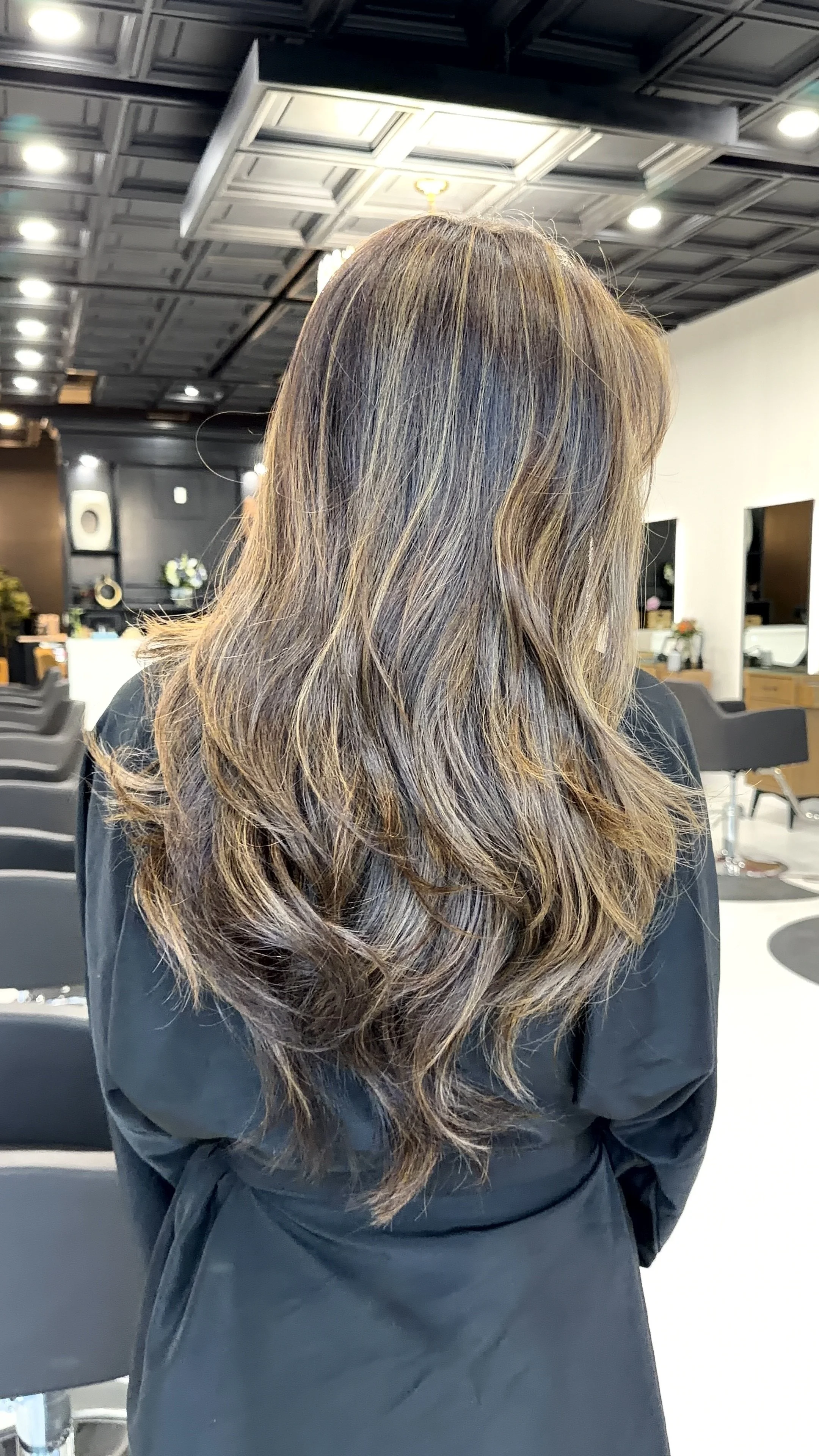 Back view of a woman with long, wavy, brown hair with highlights, sitting in a modern salon.
