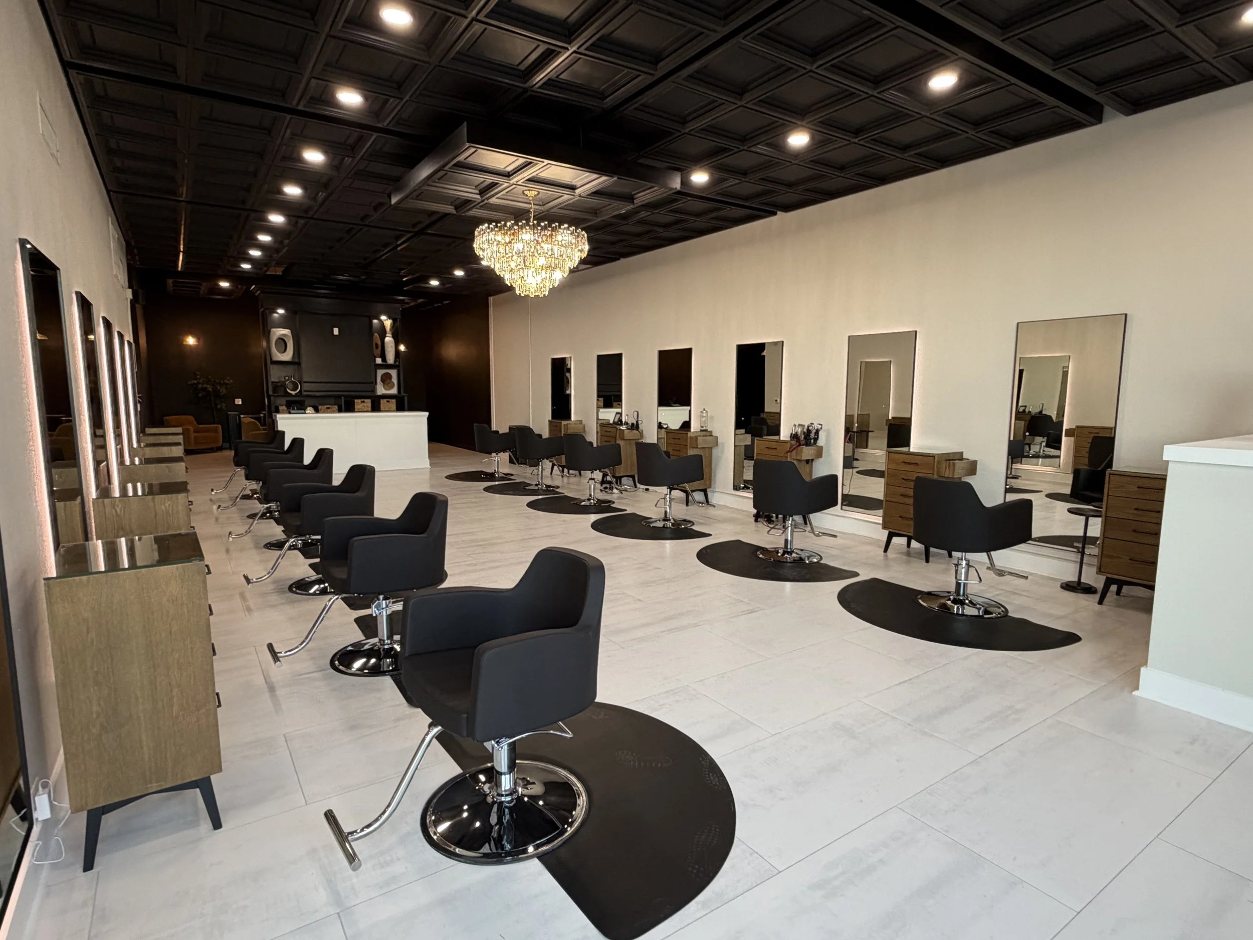 Modern hair salon with multiple styling stations, black chairs, and large mirrors, chandelier, and reception area in the background.