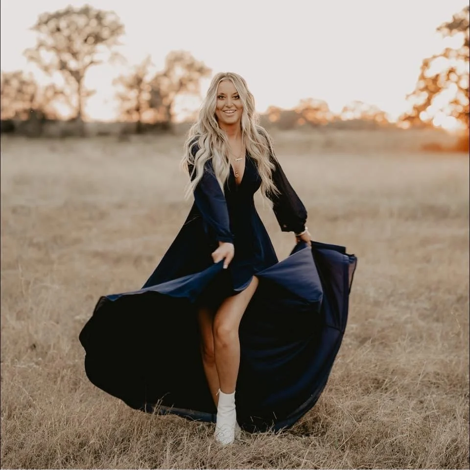 A woman with long blonde hair wearing a black dress with a deep neckline and white socks, standing in an open field during sunset.