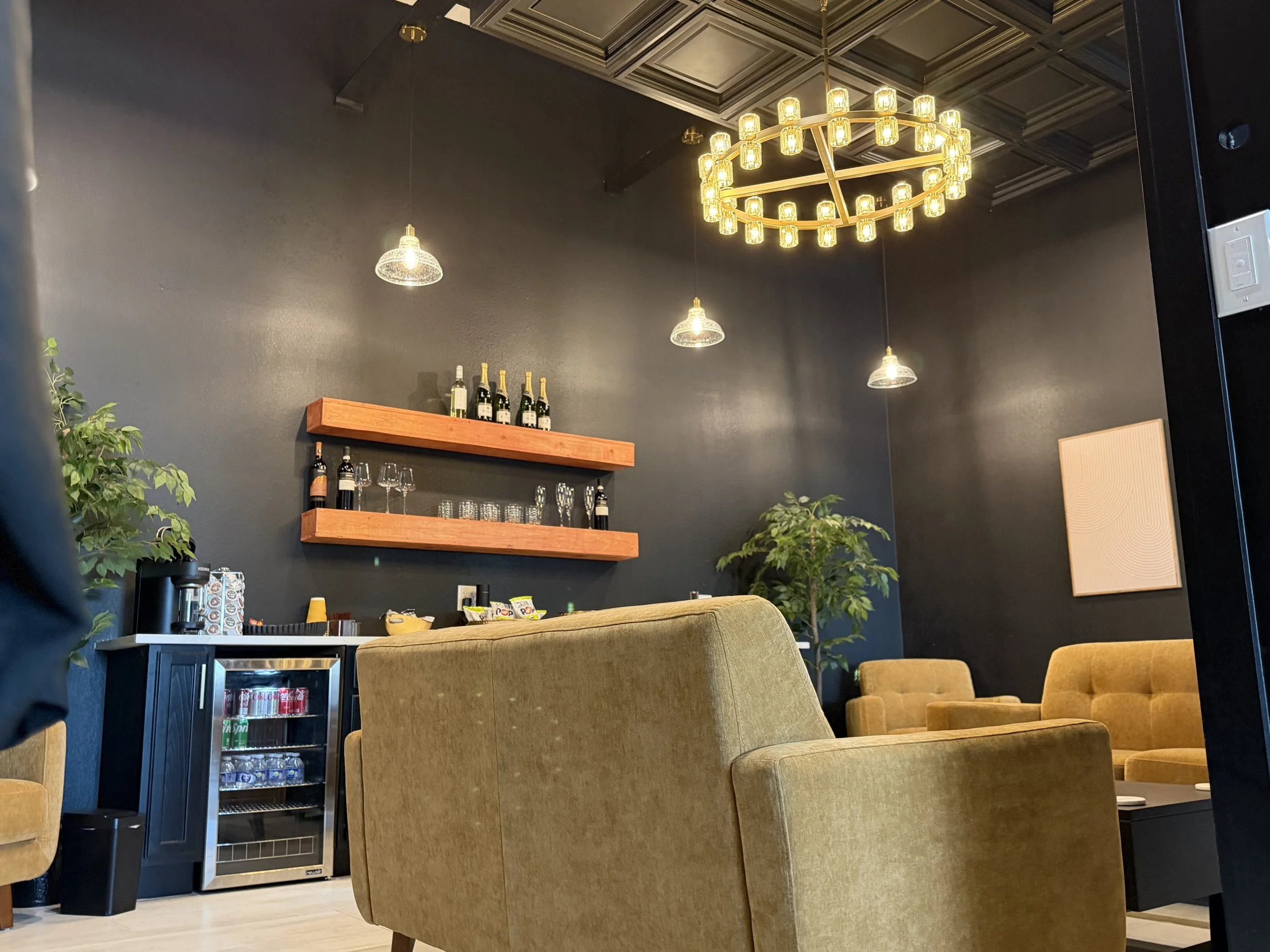 Interior of a modern lounge with yellow upholstered chairs, dark walls, and a ceiling with a circular chandelier. There are shelves with bottles and glassware, small plants, and a mini fridge.