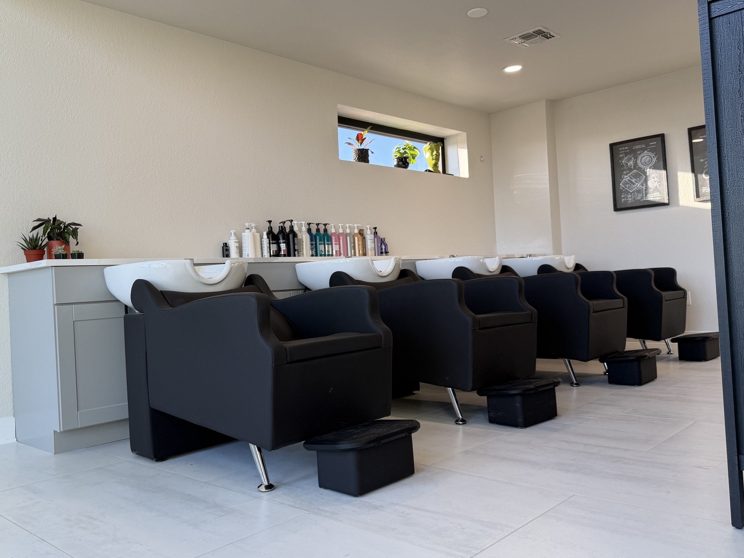 Five black salon chairs with white shampoo bowls behind them, set in a row in a bright, modern hair salon. There are bottles of shampoo and conditioner on a white counter behind the chairs, with a small potted plant and decorative items.