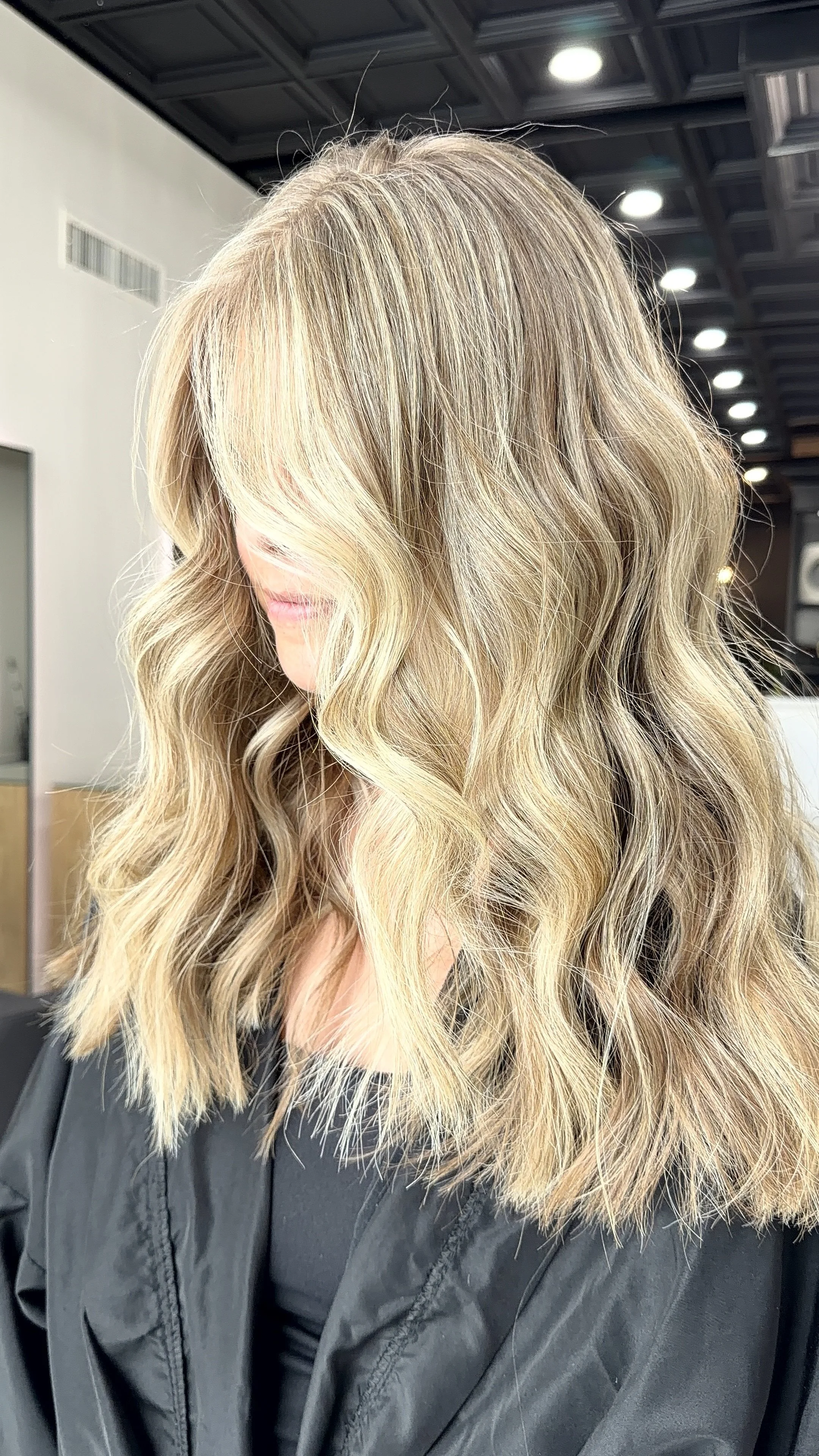 A woman with shoulder-length, wavy blonde hair styled in loose waves, wearing a black salon cape, in a salon with a ceiling featuring square patterned tiles and bright ceiling lights.