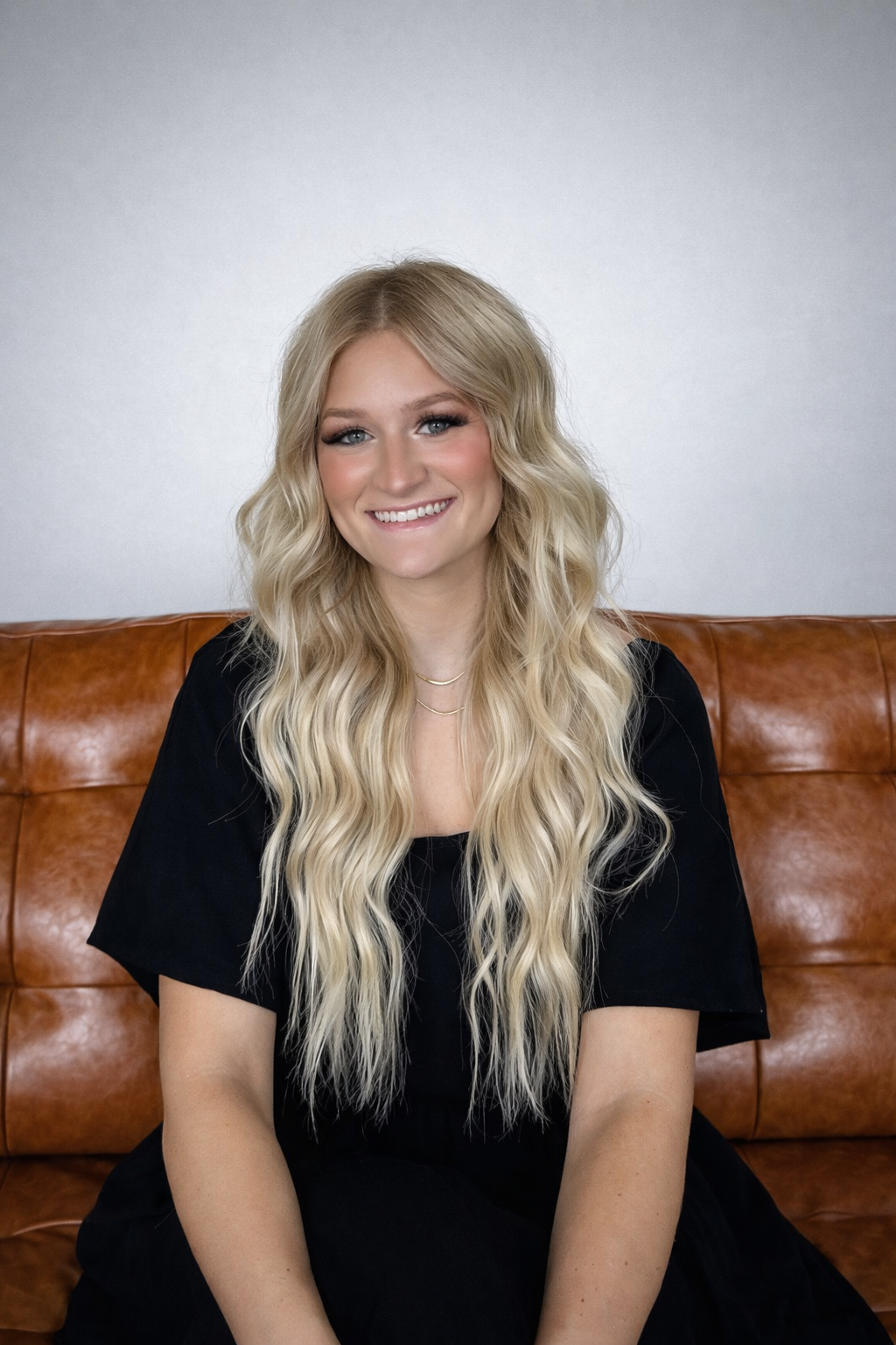 A young woman with long, wavy blonde hair and blue eyes, smiling and sitting on a brown leather couch against a plain gray wall.