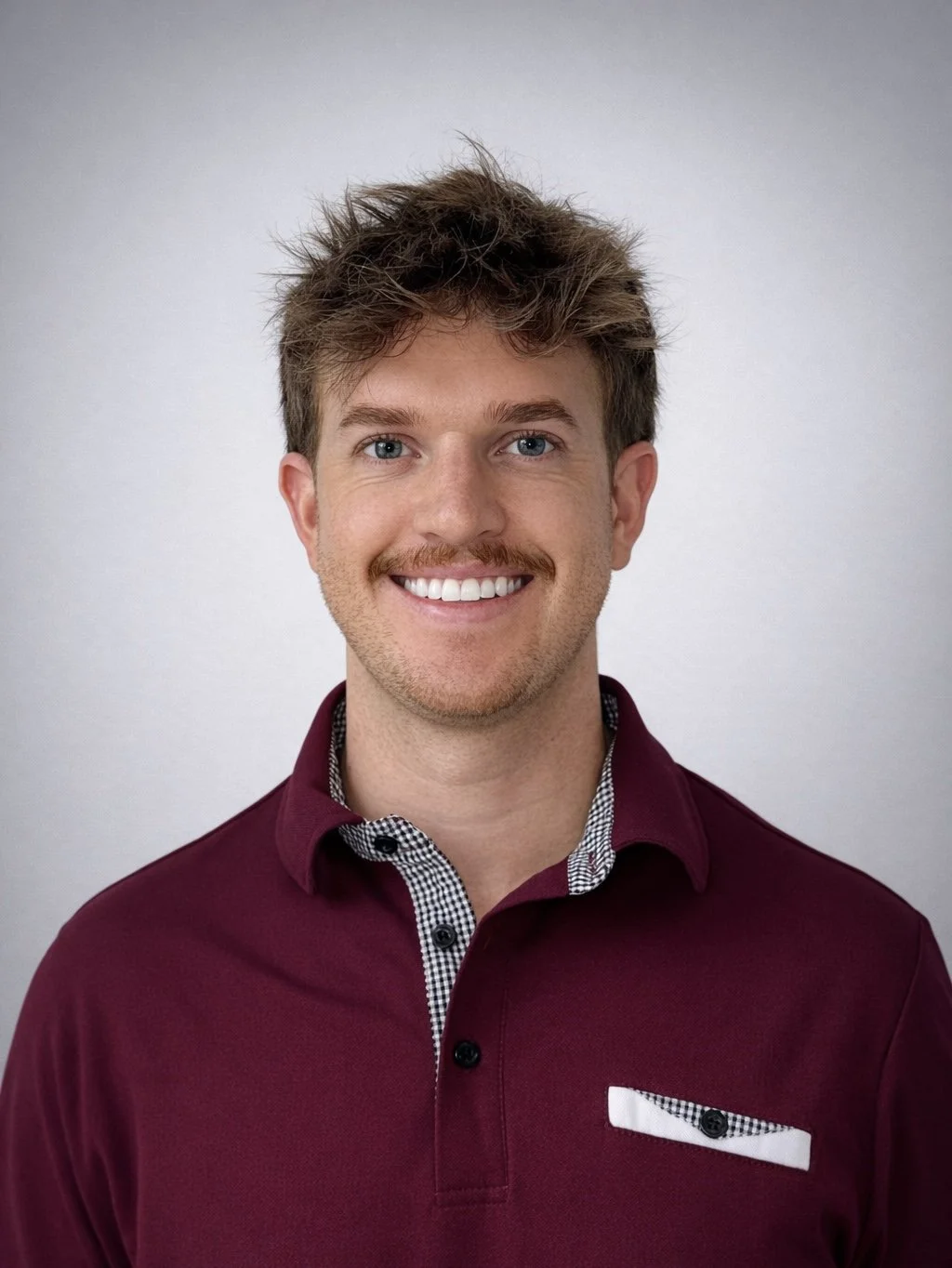 Portrait of a smiling young man with light brown hair, blue eyes, and a trimmed beard, wearing a maroon polo shirt with a checkered collar and pocket details, standing against a plain light gray background.