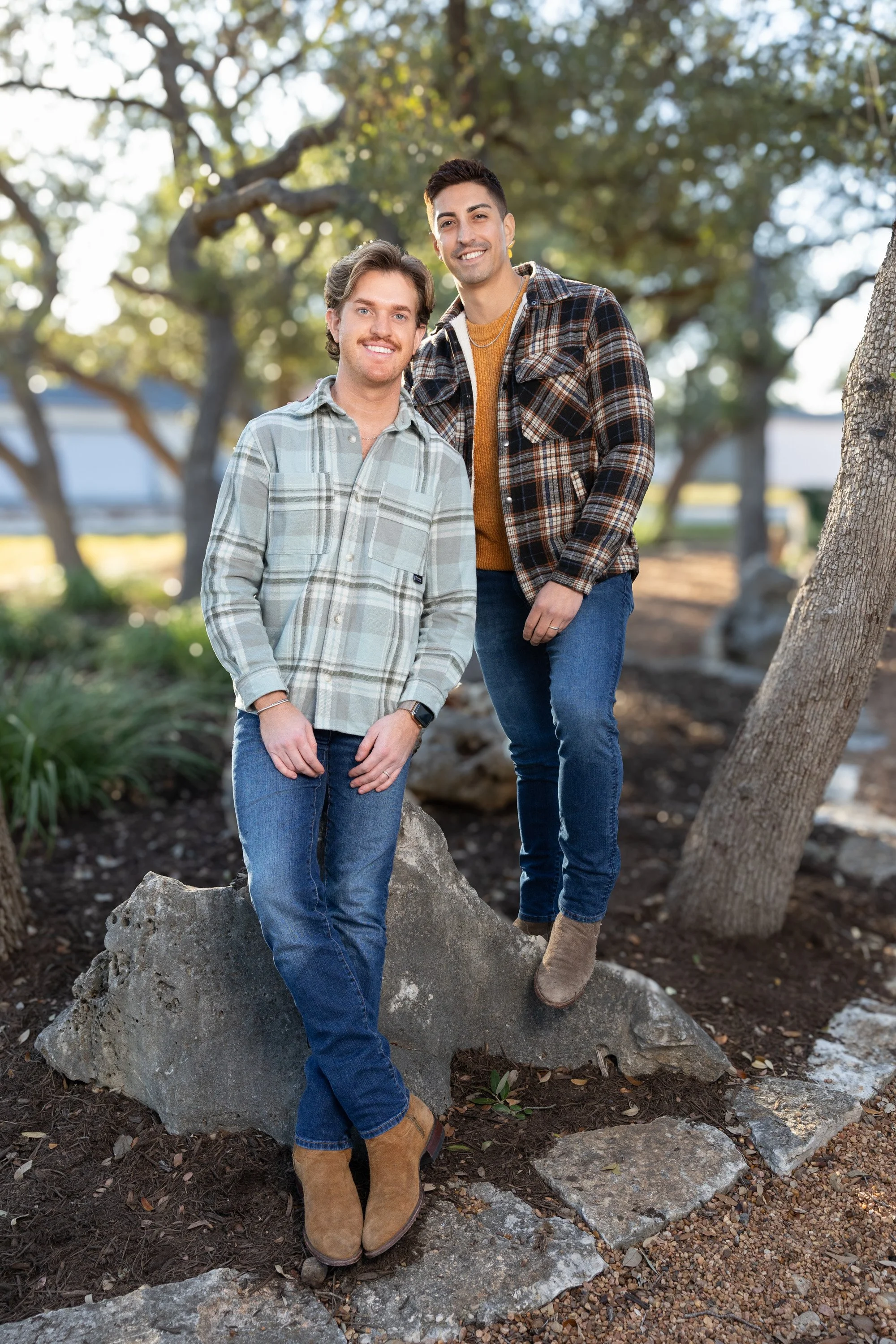 Two young men standing outdoors on rocks, smiling at the camera with trees and sunlight in the background.