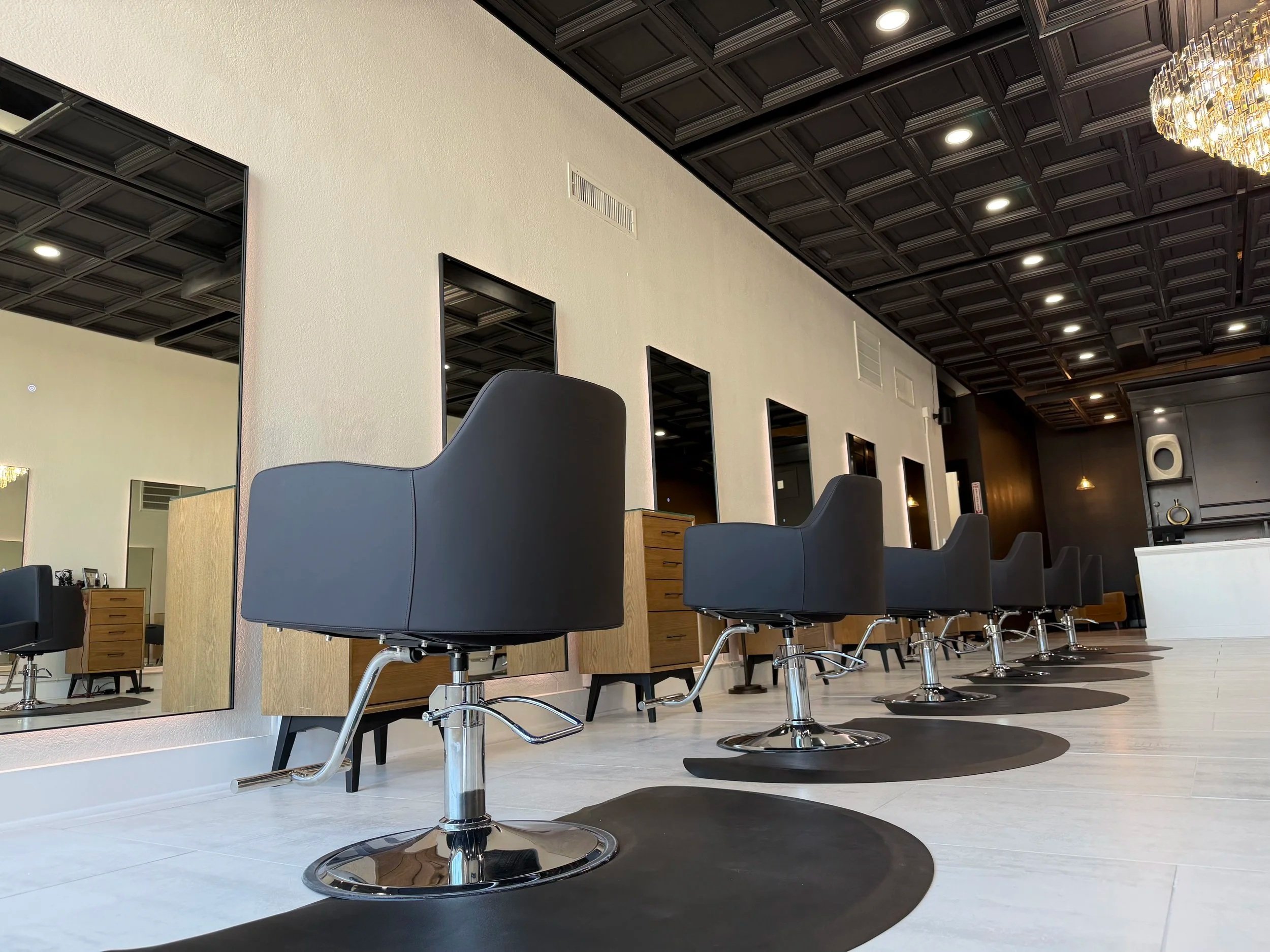 A modern hair salon with black styling chairs and large mirrors on the wall. The salon has a dark coffered ceiling, floor mirrors, and minimal decor.