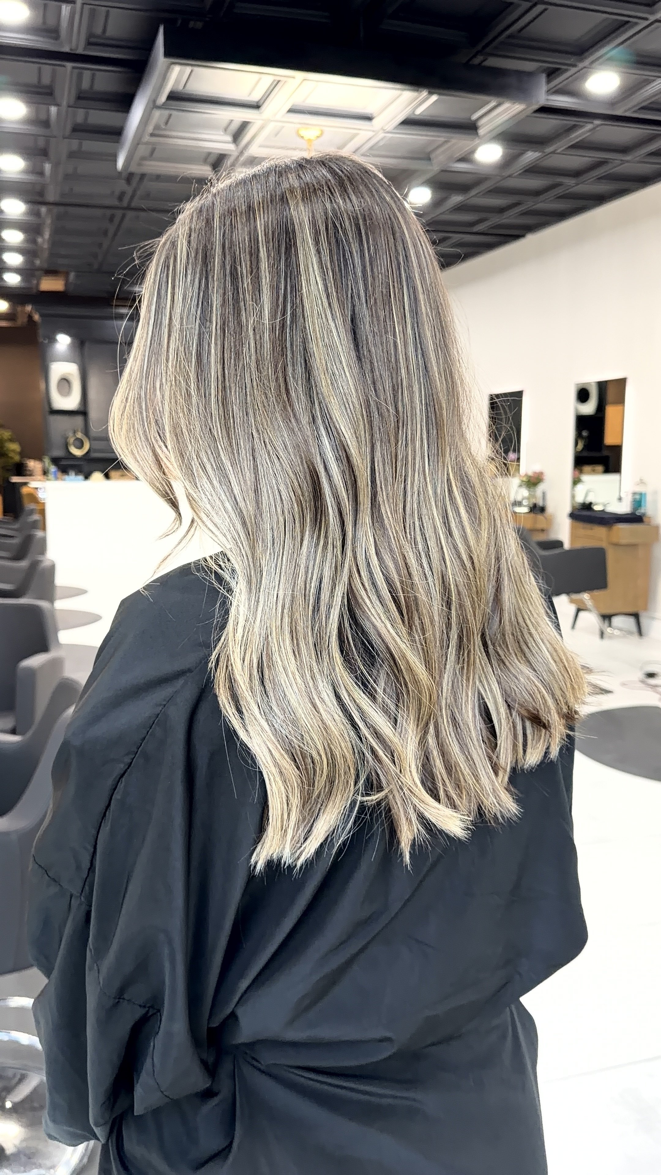 Side view of a woman with shoulder-length wavy hair with blonde highlights, sitting in a hair salon.