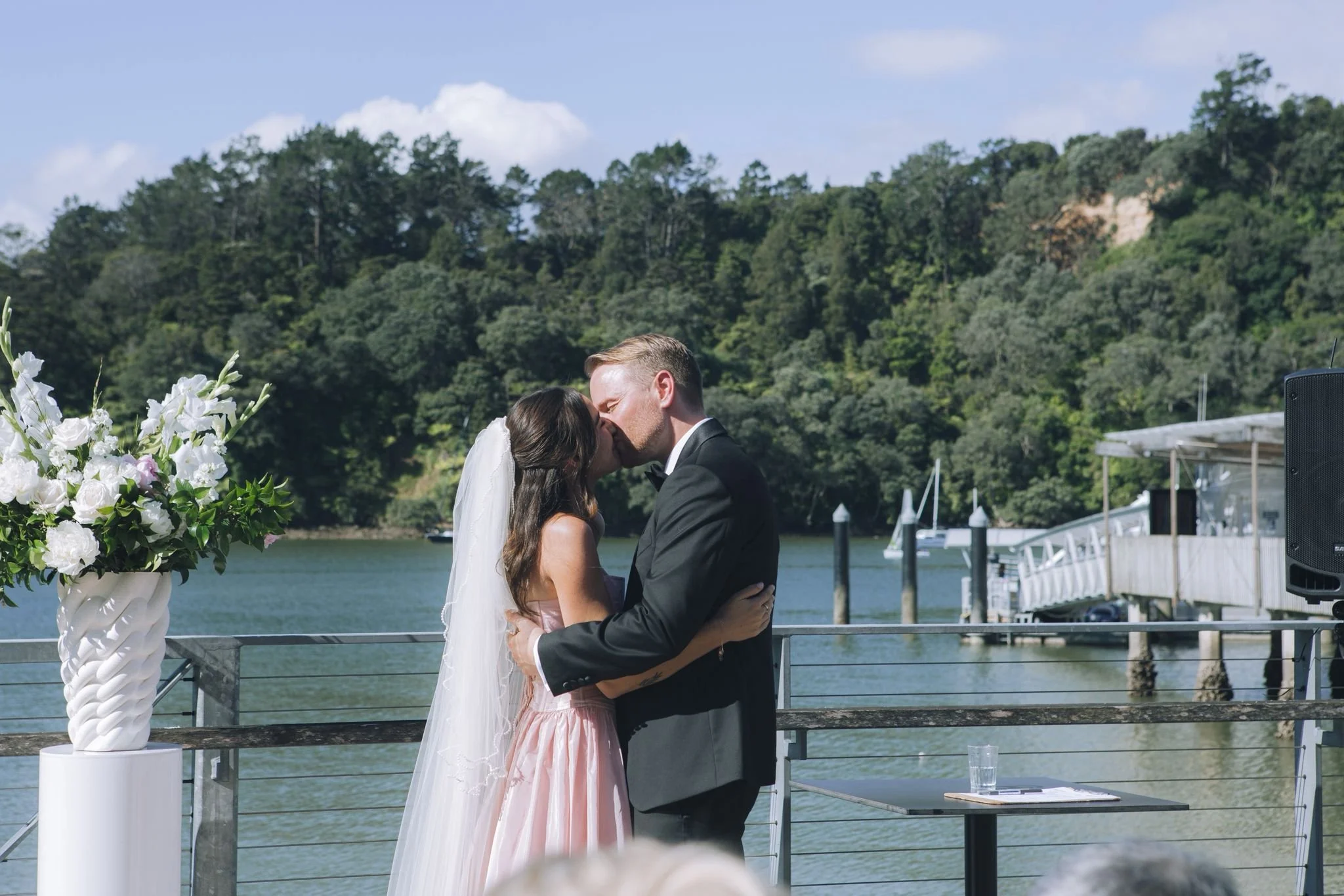 A bride and groom kissing during their outdoor wedding ceremony by the water, with a bouquet of white flowers on a stand nearby and lush green trees in the background.